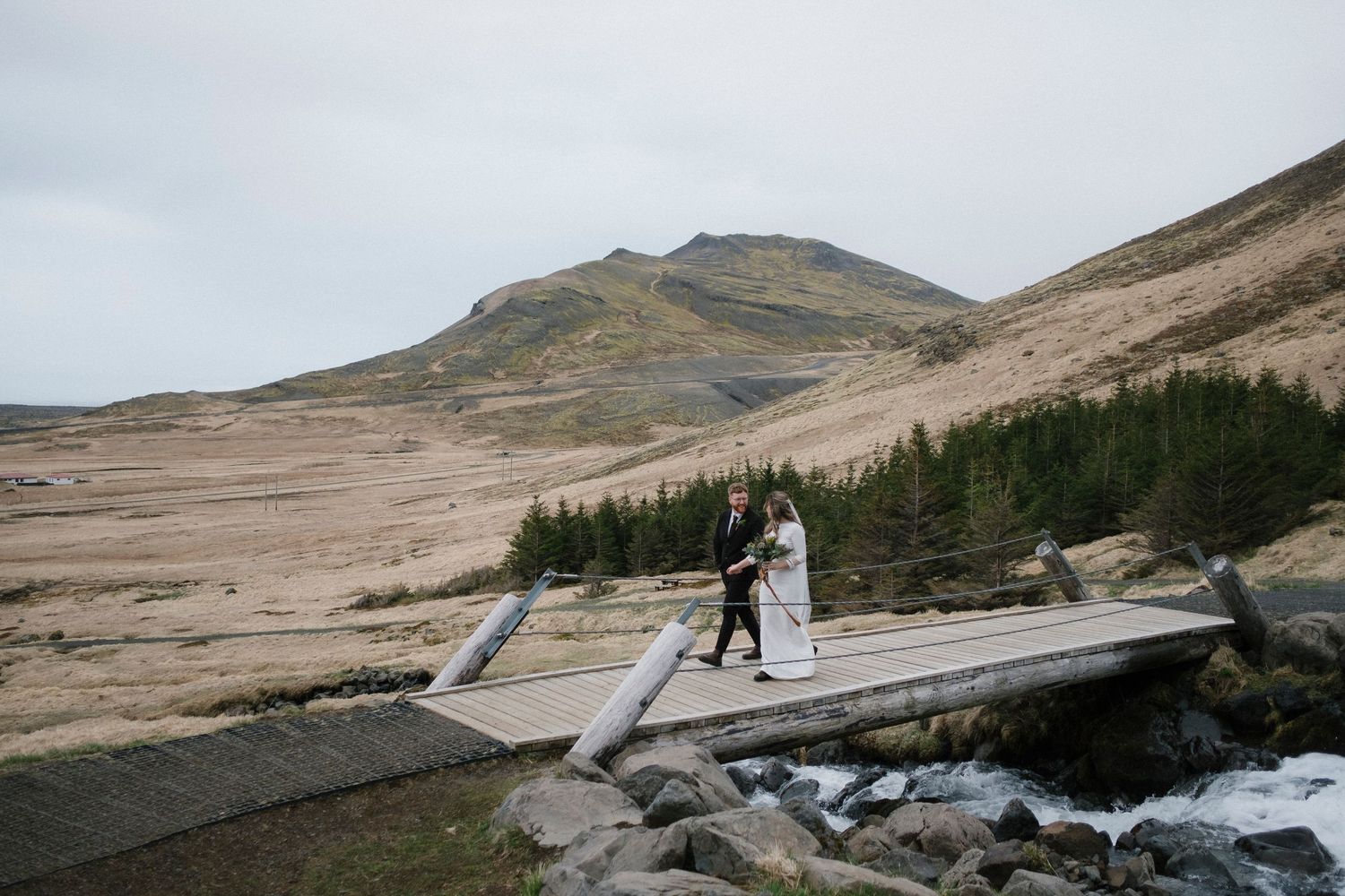 A couple walks across a wooden bridge over a stream with mountains in the background.