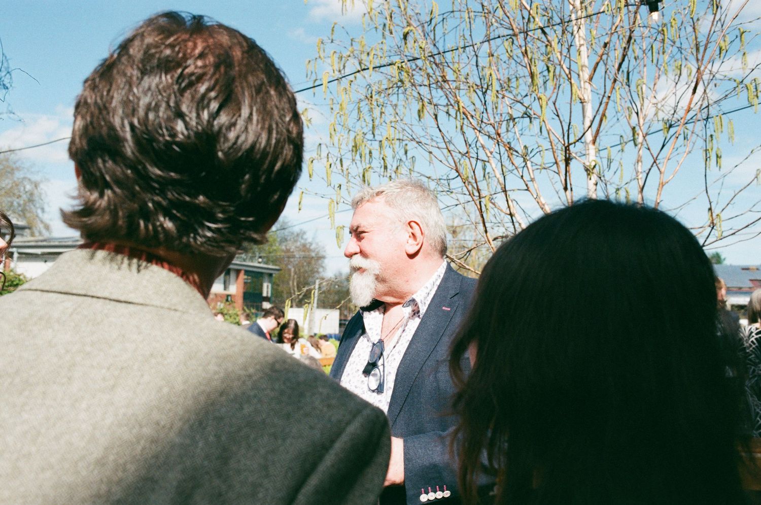 Man in blue blazer speaking outdoors with others, trees visible in sunny background.