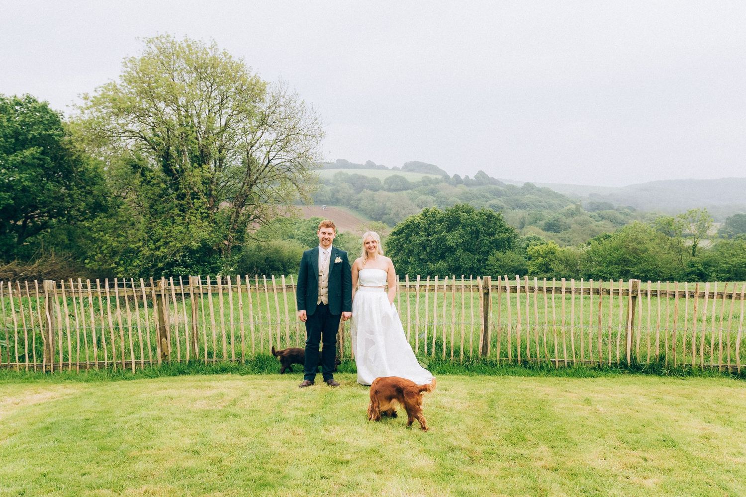 Bride and groom stand in green garden with a golden retriever dog and rustic wooden fence.