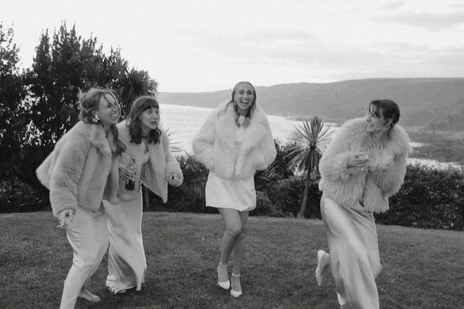 Four women in white outfits and fur coats laughing outdoors with scenic water views.
