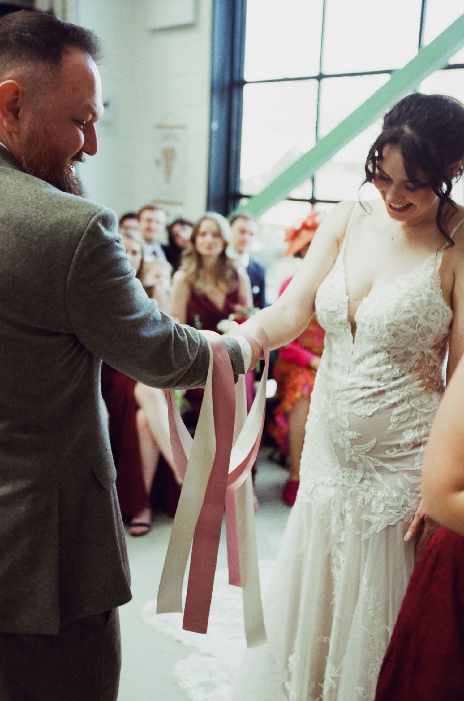 Bride in lace gown and groom in gray suit perform handfasting ceremony with colorful ribbons, guests watching in background.