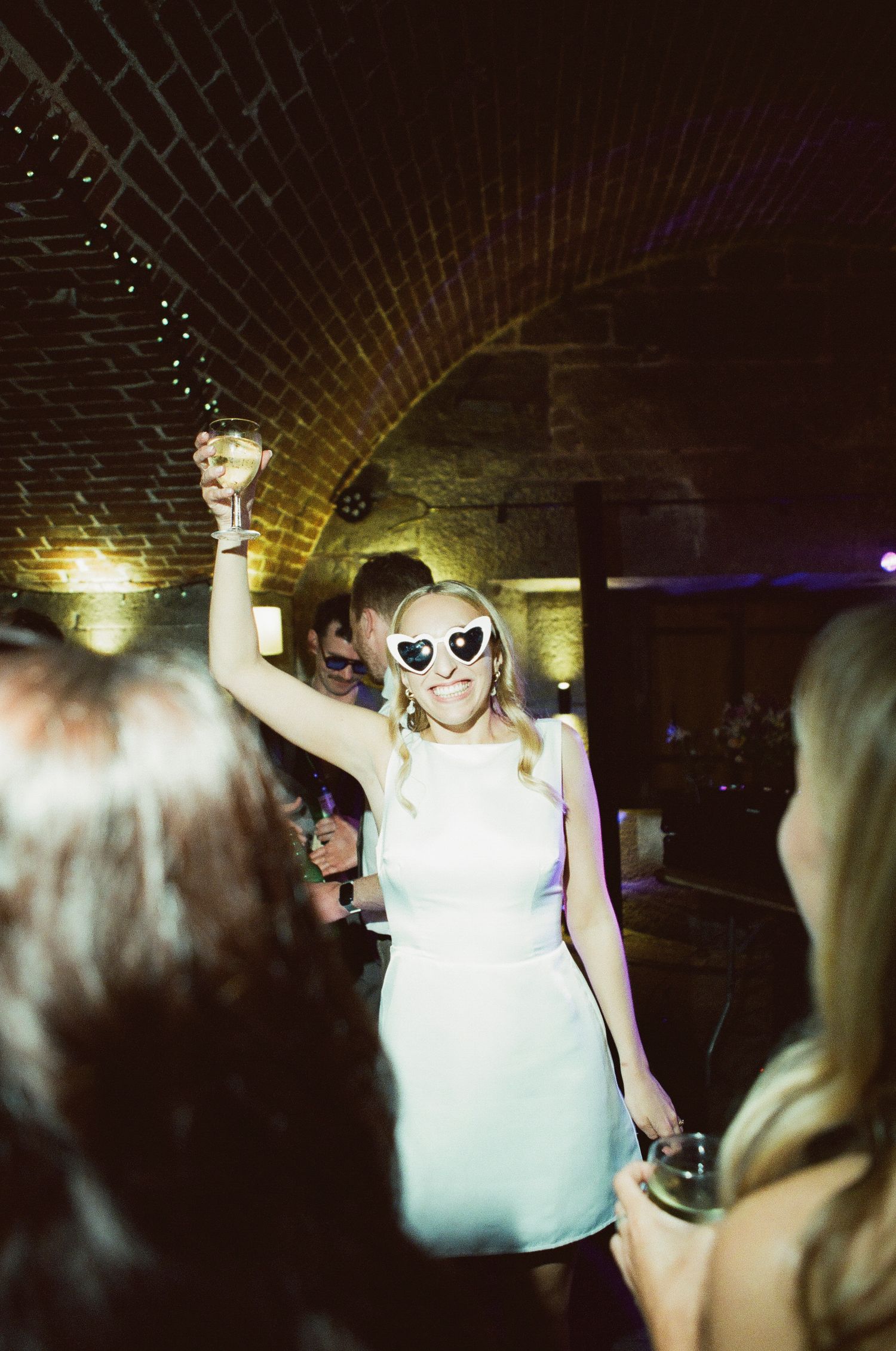 Bride dancing joyfully in white mini dress wearing heart-shaped sunglasses at wedding reception.
