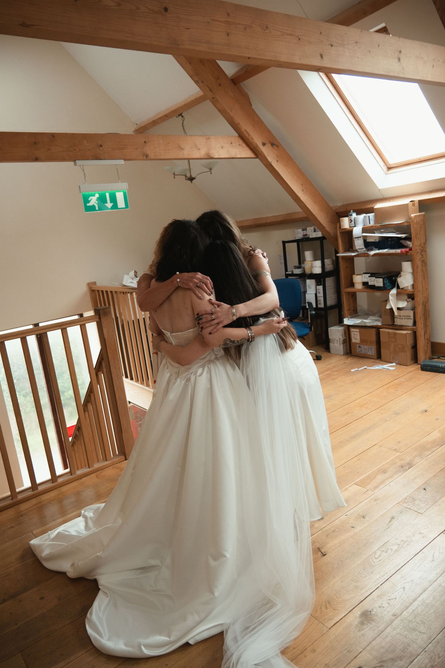 Two brides sharing an emotional hug in a rustic barn venue with wooden beams and hardwood floors.