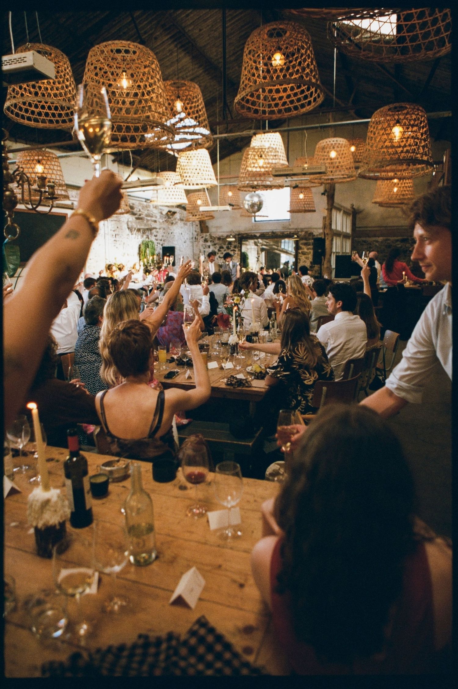 Wedding reception guests raising glasses in a toast inside a rustic venue with wicker pendant lights.