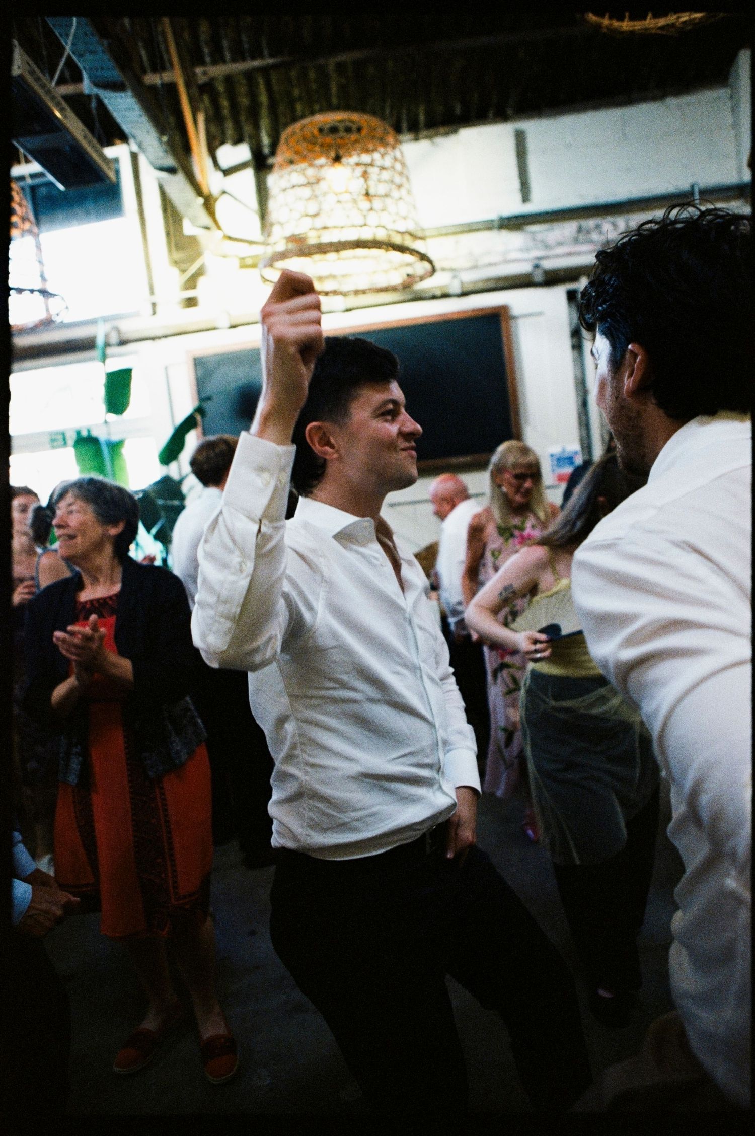 Man in white shirt dancing joyfully at an indoor party with guests and a chandelier in the background.