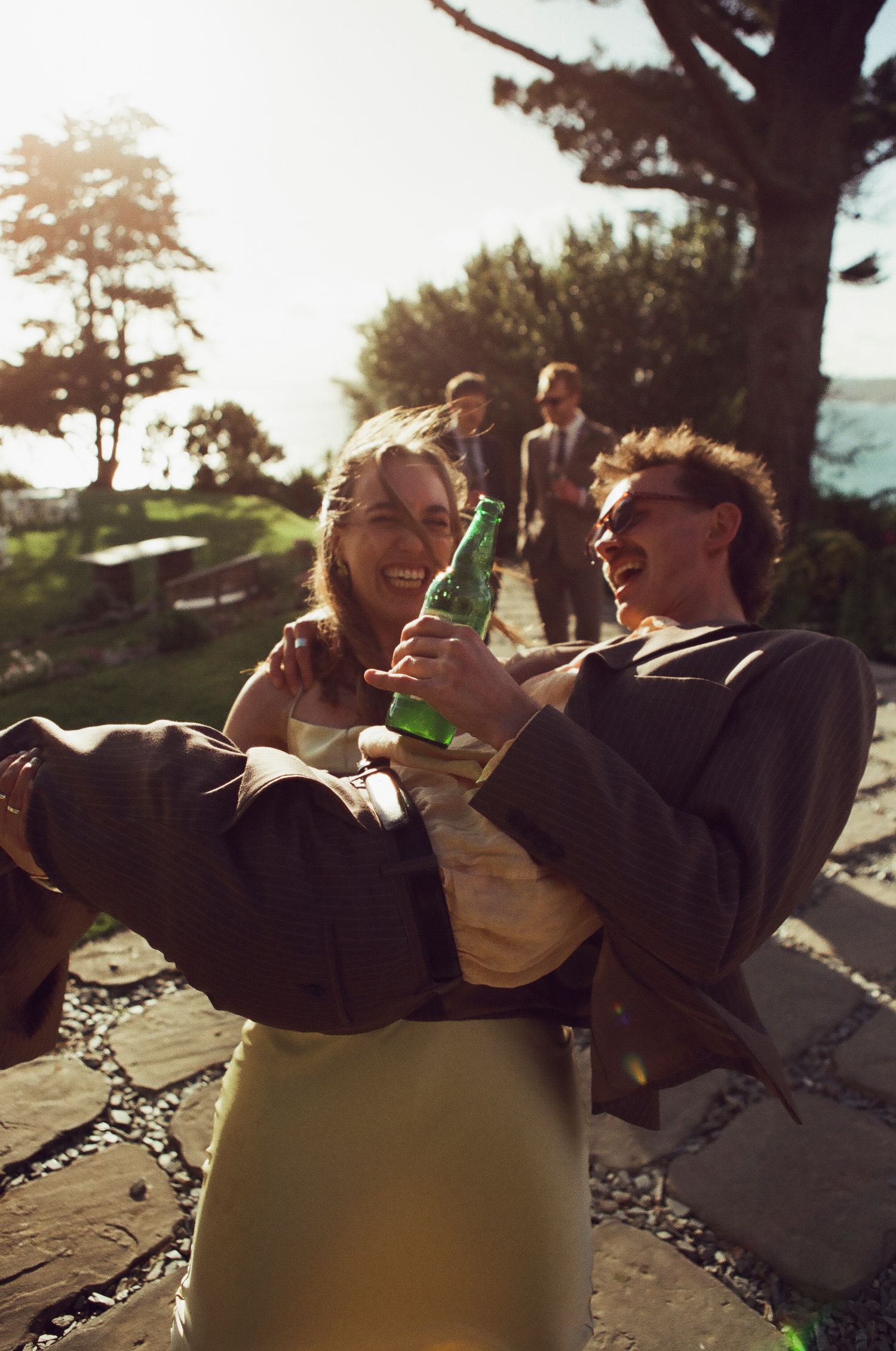 Two people laughing and toasting with green beer bottles outdoors at a sunny coastal gathering.
