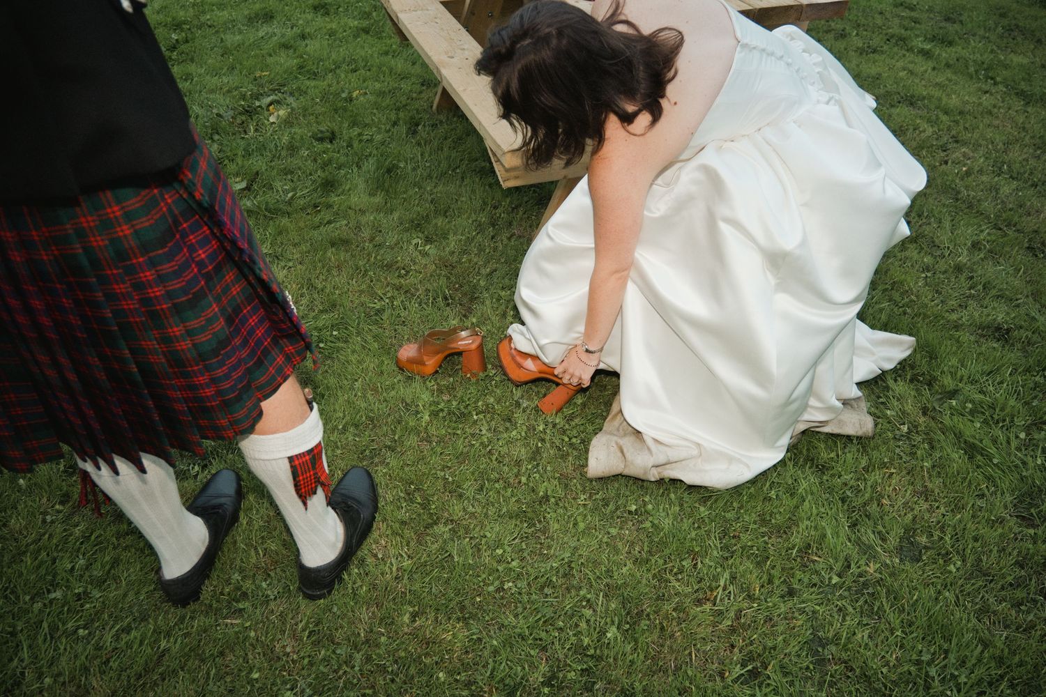 Bride in white gown adjusting brown sandals beside groom wearing traditional Scottish tartan kilt on green grass.