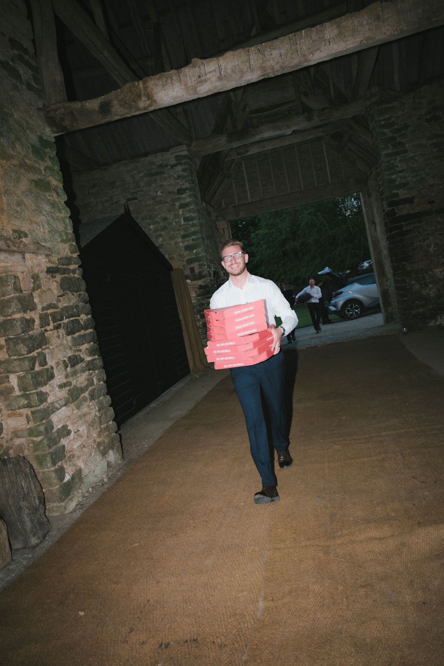 Man in white shirt carrying stack of red pizza boxes through rustic stone barn venue at evening event.