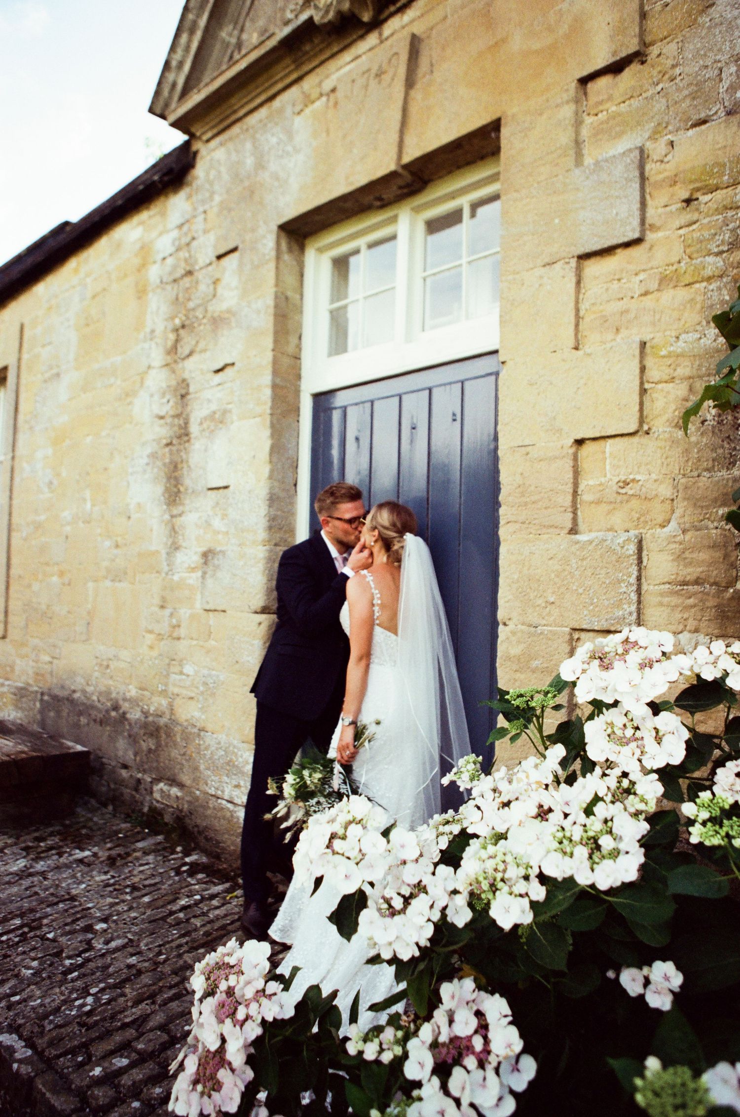 A bride and groom share a romantic kiss by a stone building with a blue door, surrounded by white hydrangeas.