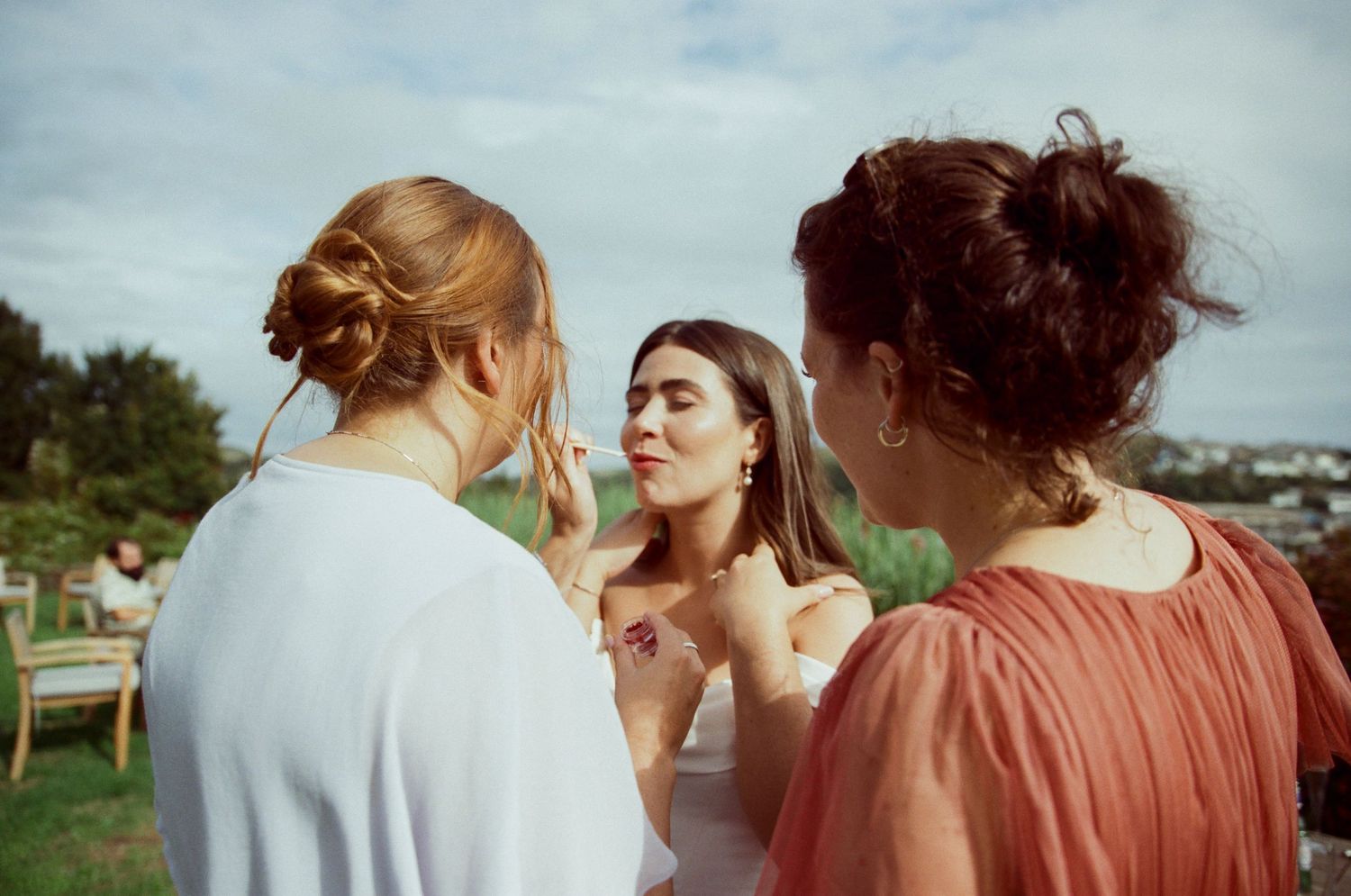 Three women in elegant dresses gather outdoors at a garden event, engaged in conversation under a cloudy sky.