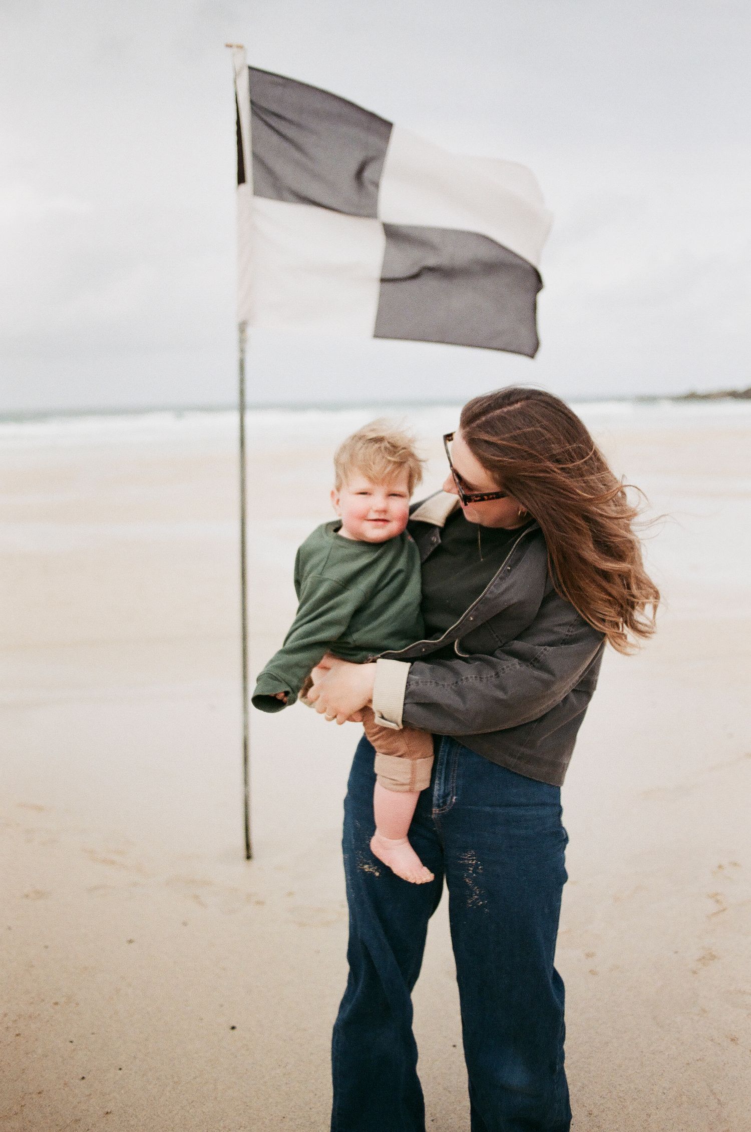 A mother holds her young child on a sandy beach with a black and white checkered flag behind them.