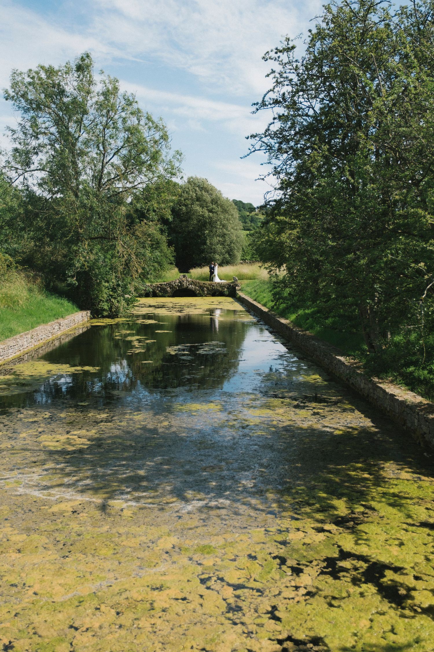 A serene canal lined with stone walls and algae-covered water, surrounded by lush green trees under a partly cloudy sky.