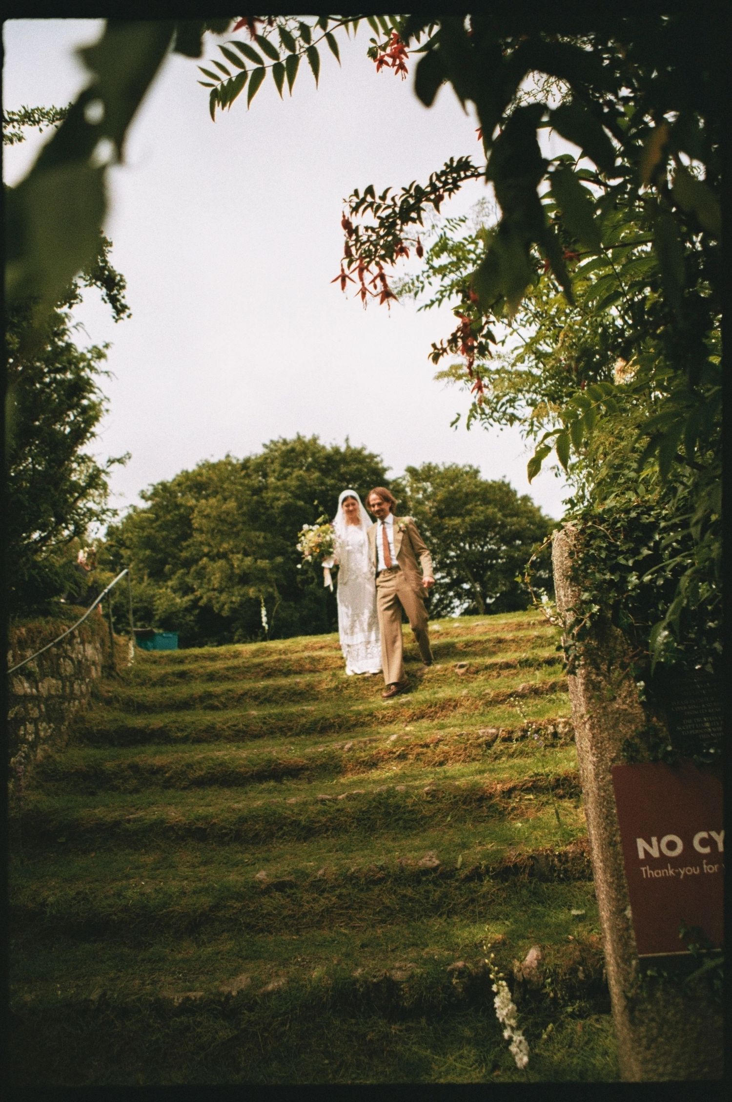 Bride and groom walking down grassy terraced steps surrounded by lush green trees.