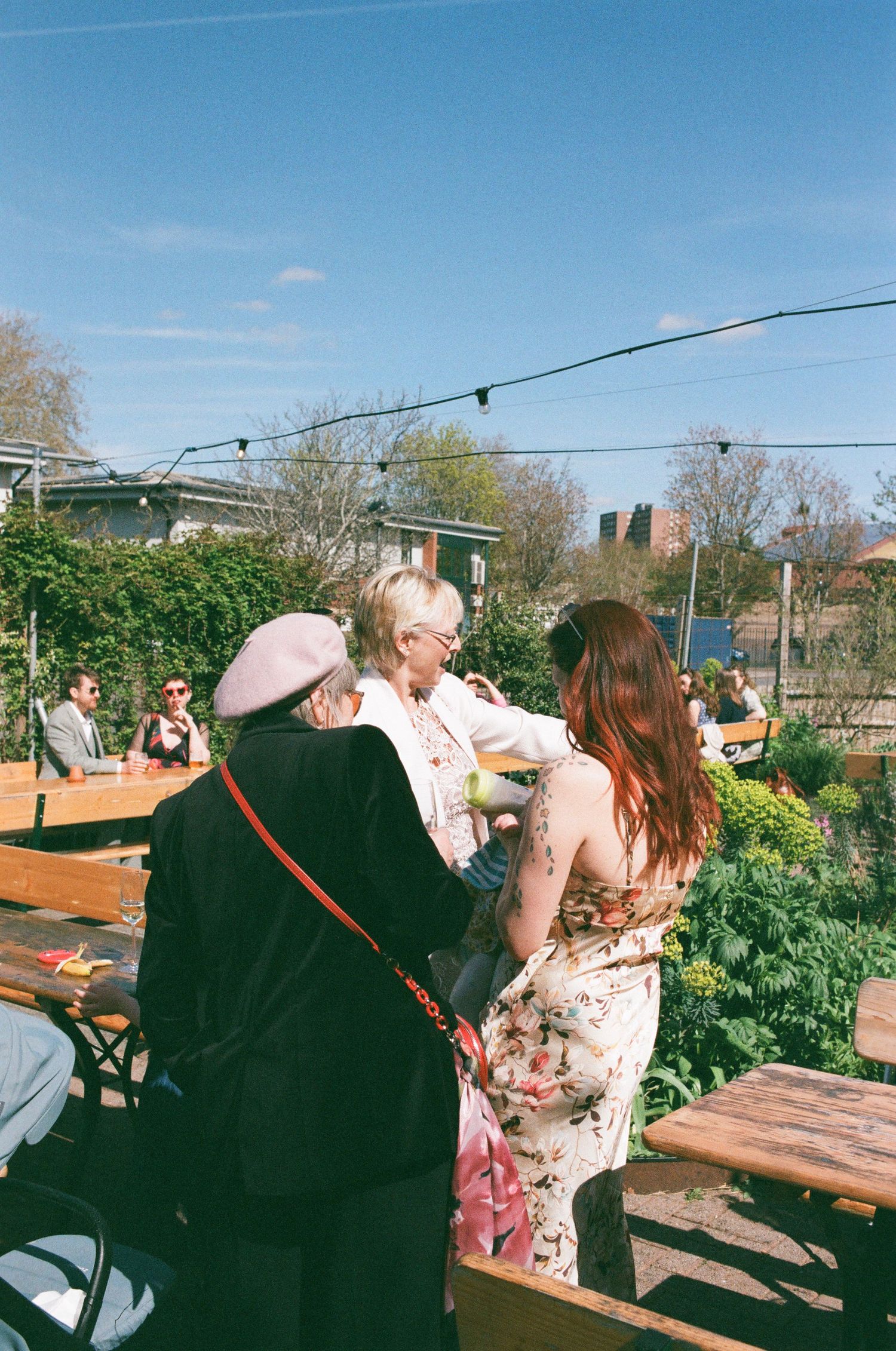 Three women socializing at an outdoor beer garden on a sunny day, surrounded by wooden benches and string lights.