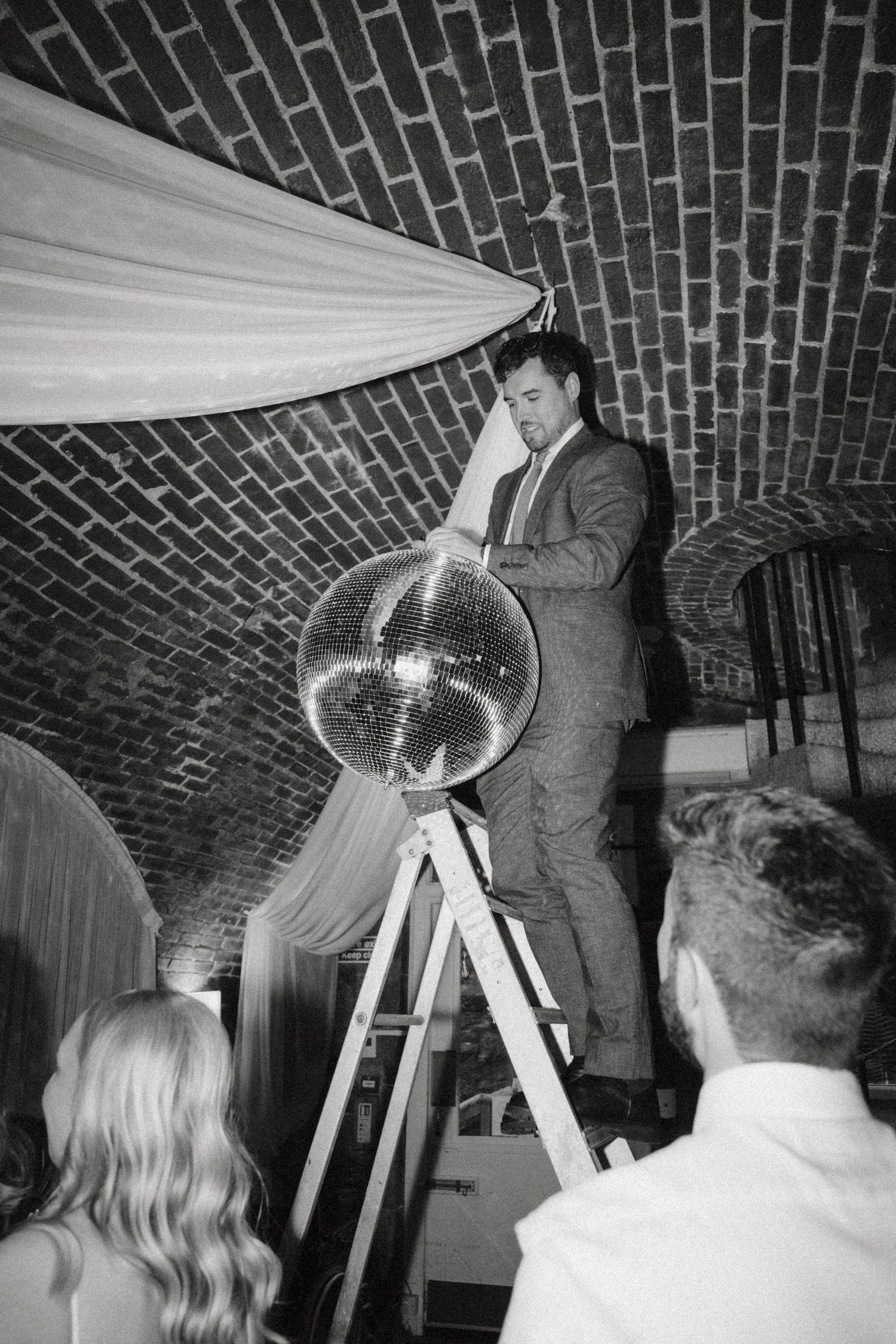 Groom on a ladder installing a large disco ball in a brick-vaulted wedding reception venue.