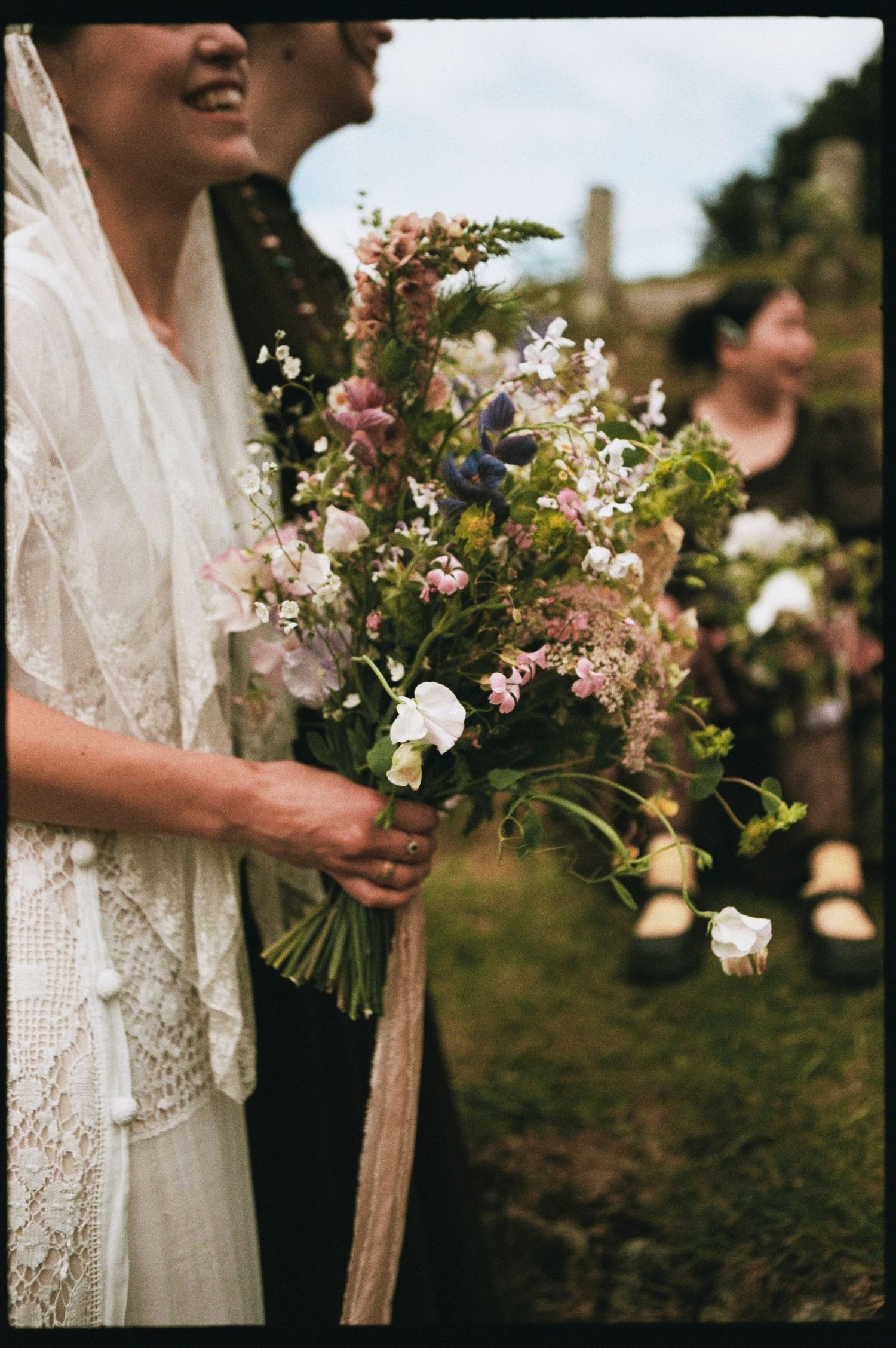 Bride in lace dress holding a wildflower bouquet with blue and white blooms at an outdoor wedding ceremony.