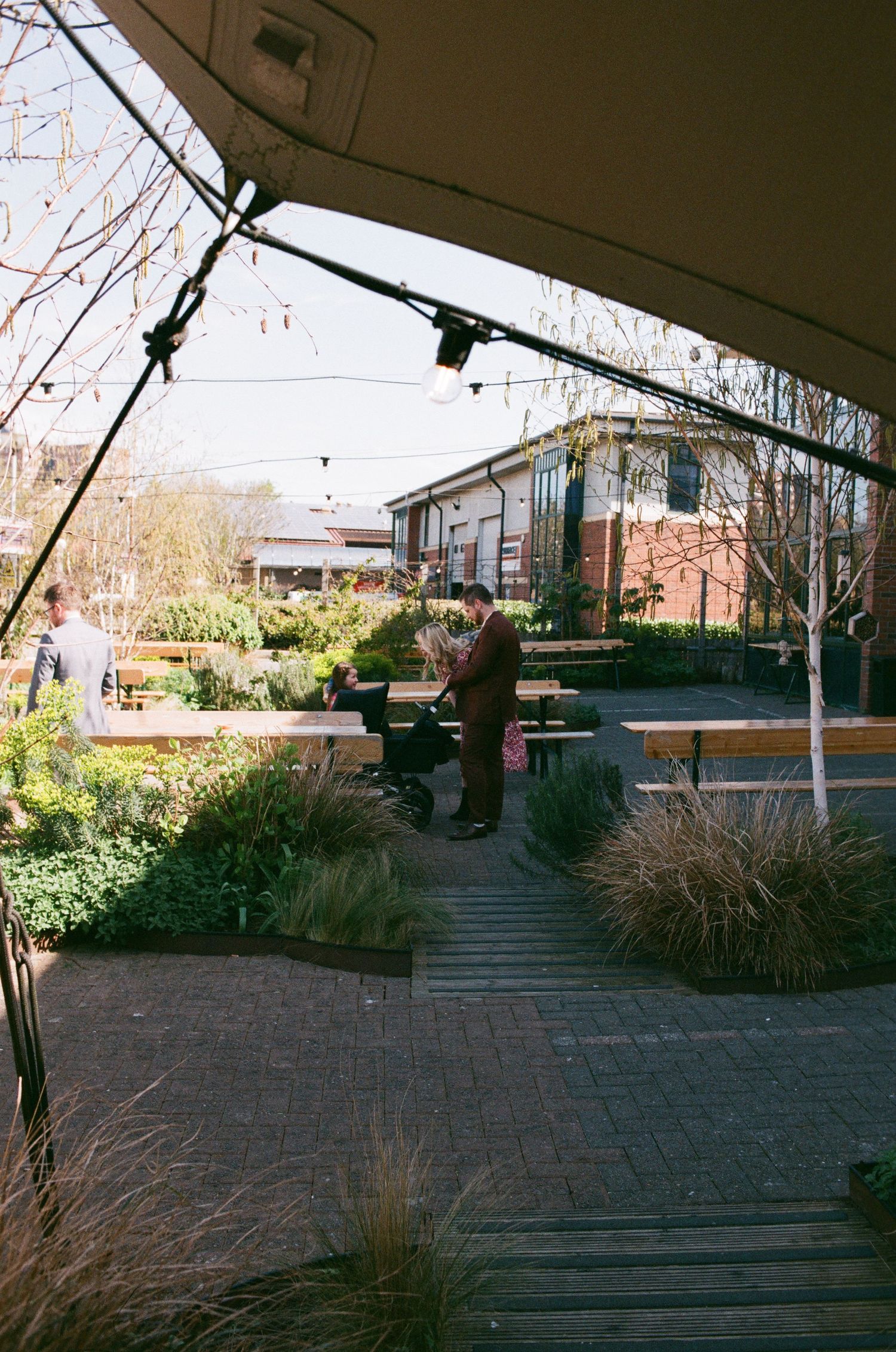 Outdoor garden cafe with wooden pathways, lush greenery, string lights, and people dining under a canopy on a sunny day.