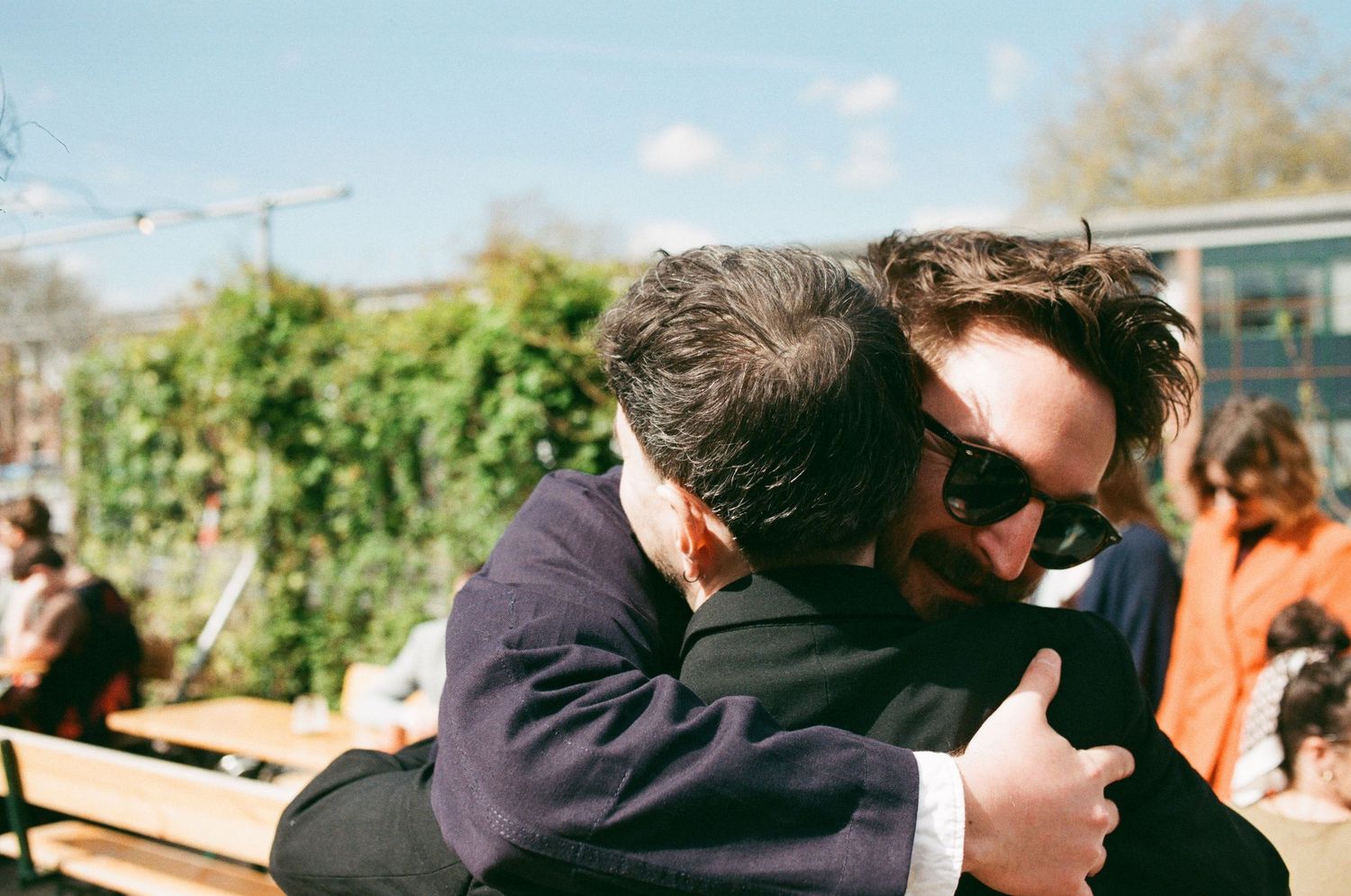 Two men in suits share a warm embrace outdoors on a sunny day at a casual gathering.