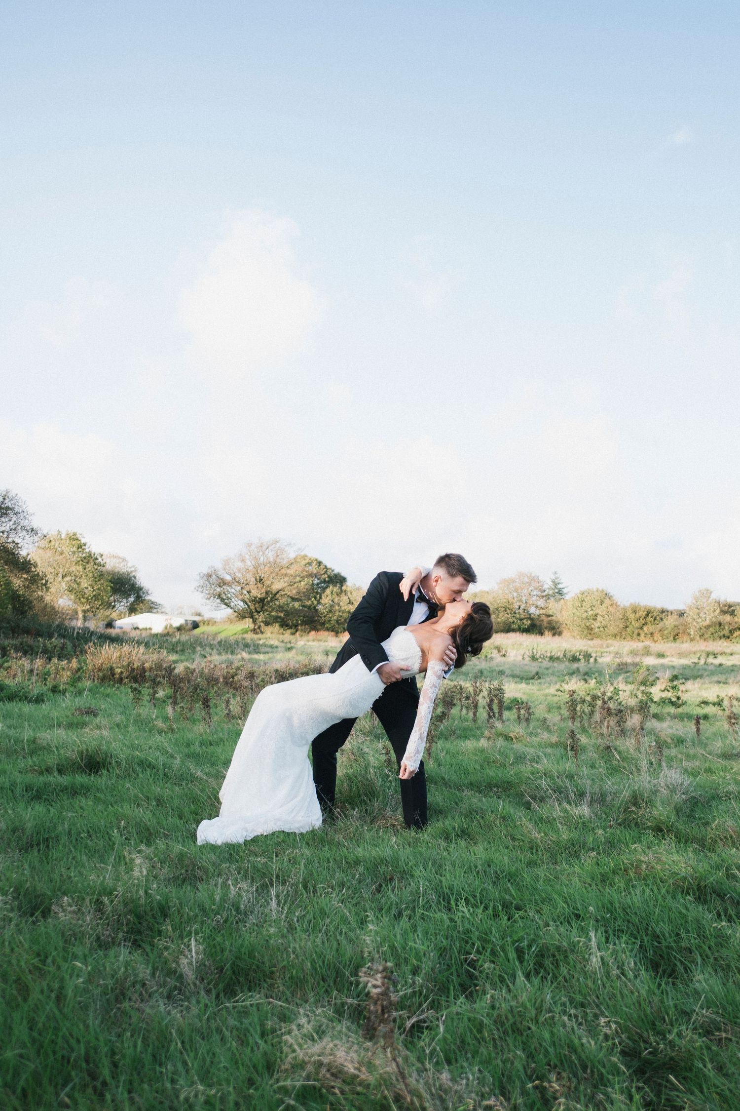Groom dips bride for a romantic kiss in a lush green field under a bright blue sky.