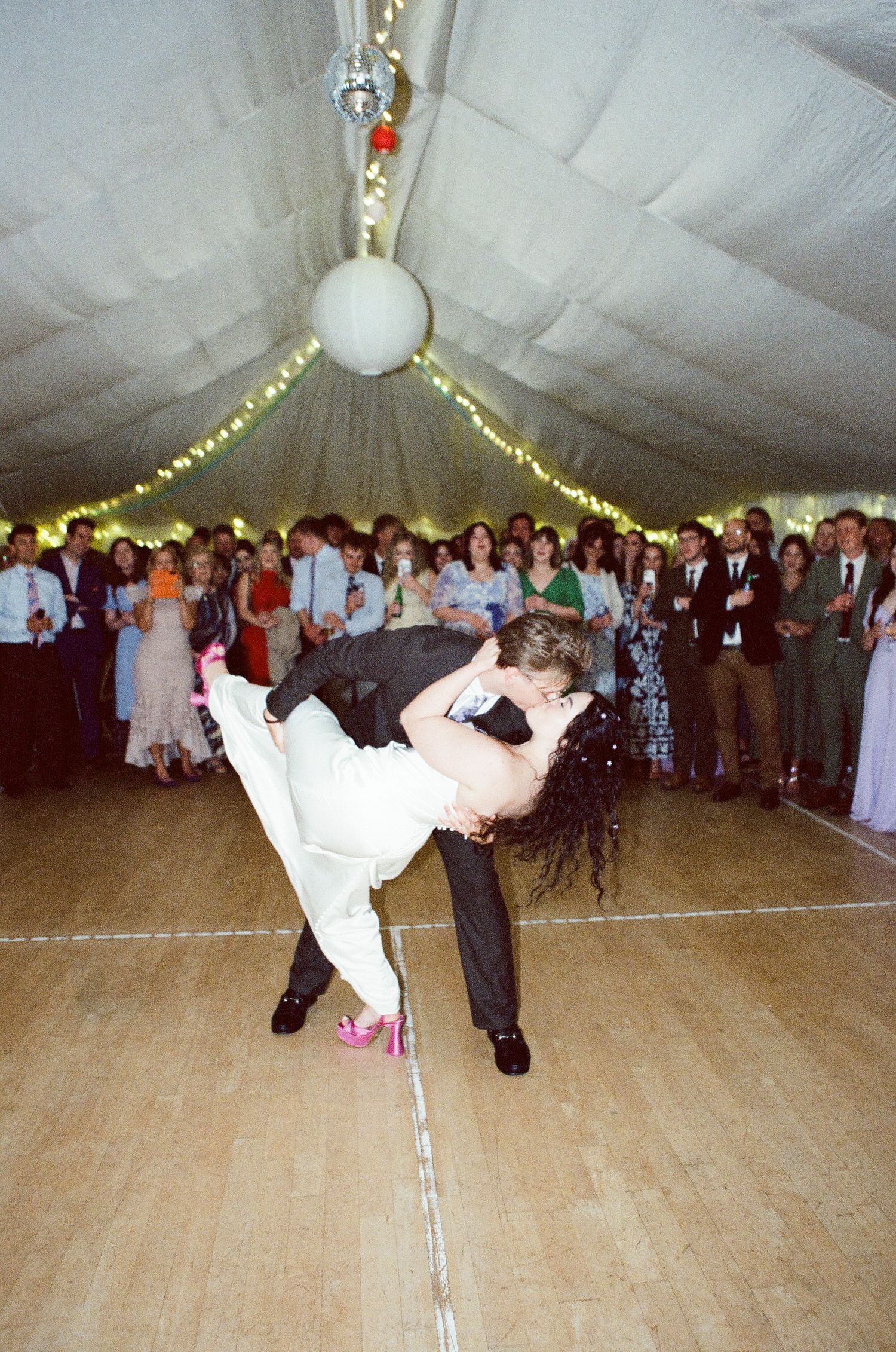 Bride and groom share a romantic dip kiss on the dance floor during their first wedding dance, surrounded by guests.