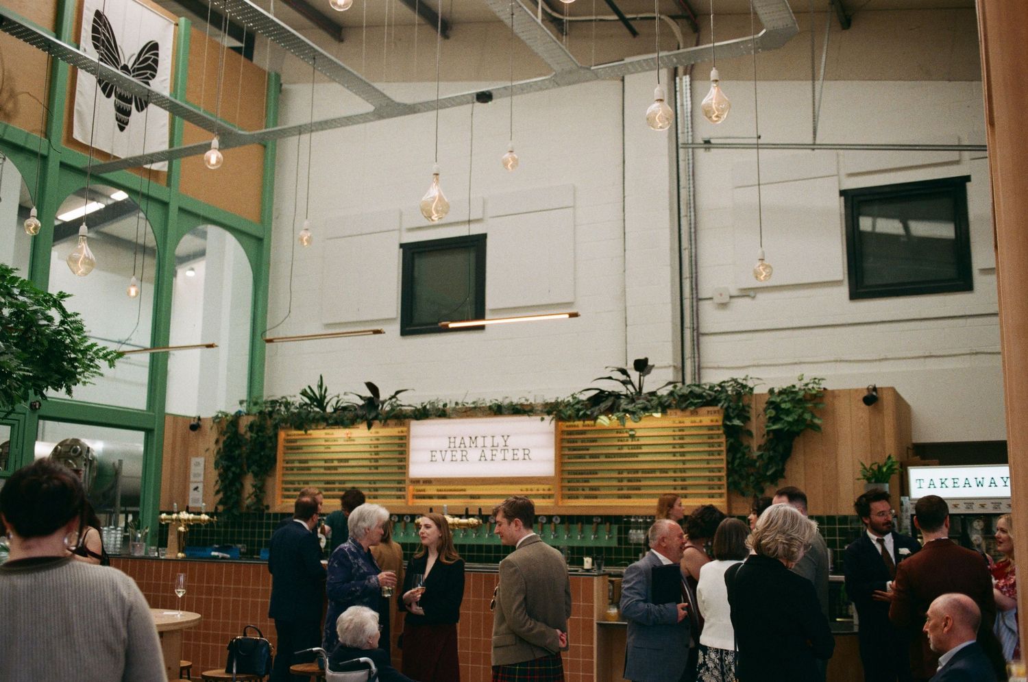 People mingling inside a modern industrial venue with hanging lights, green plants, and a wooden bar backdrop.