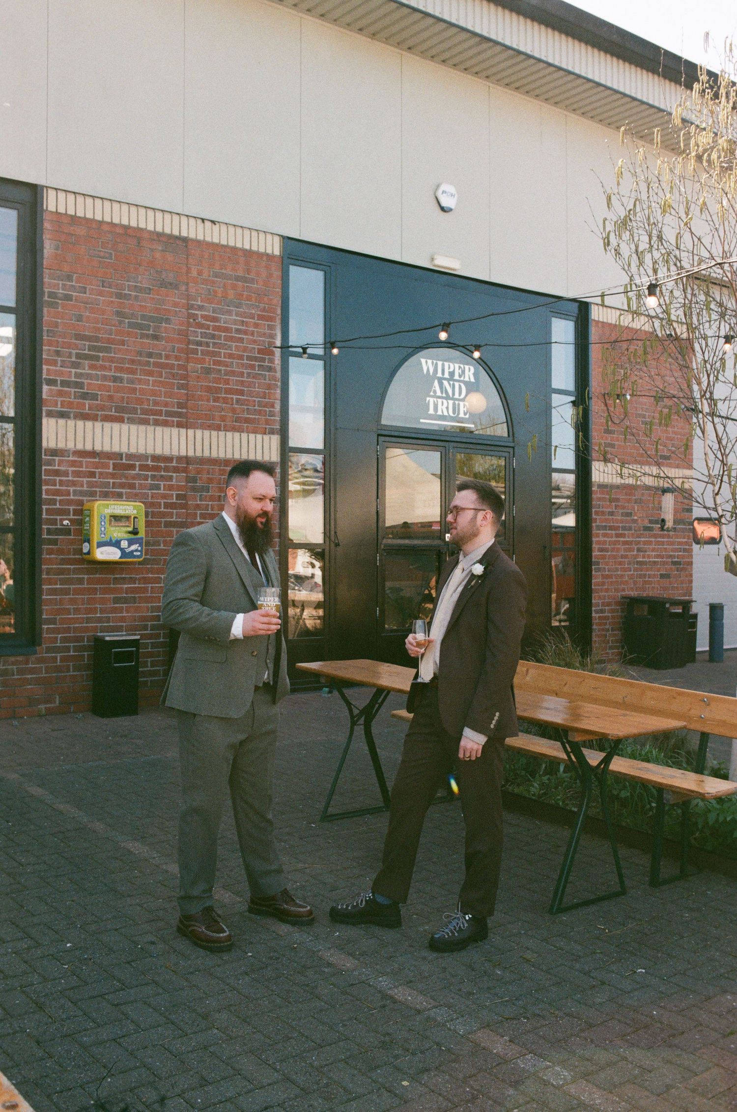Two men in suits chat outside Wiper and True brewery, holding drinks on a sunny day.