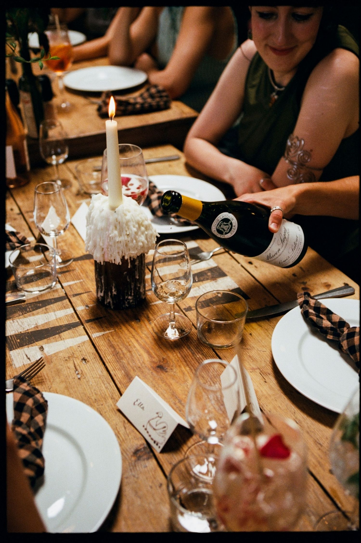 Woman pouring wine at a rustic wooden wedding reception table with candles, white plates, and floral centerpieces.