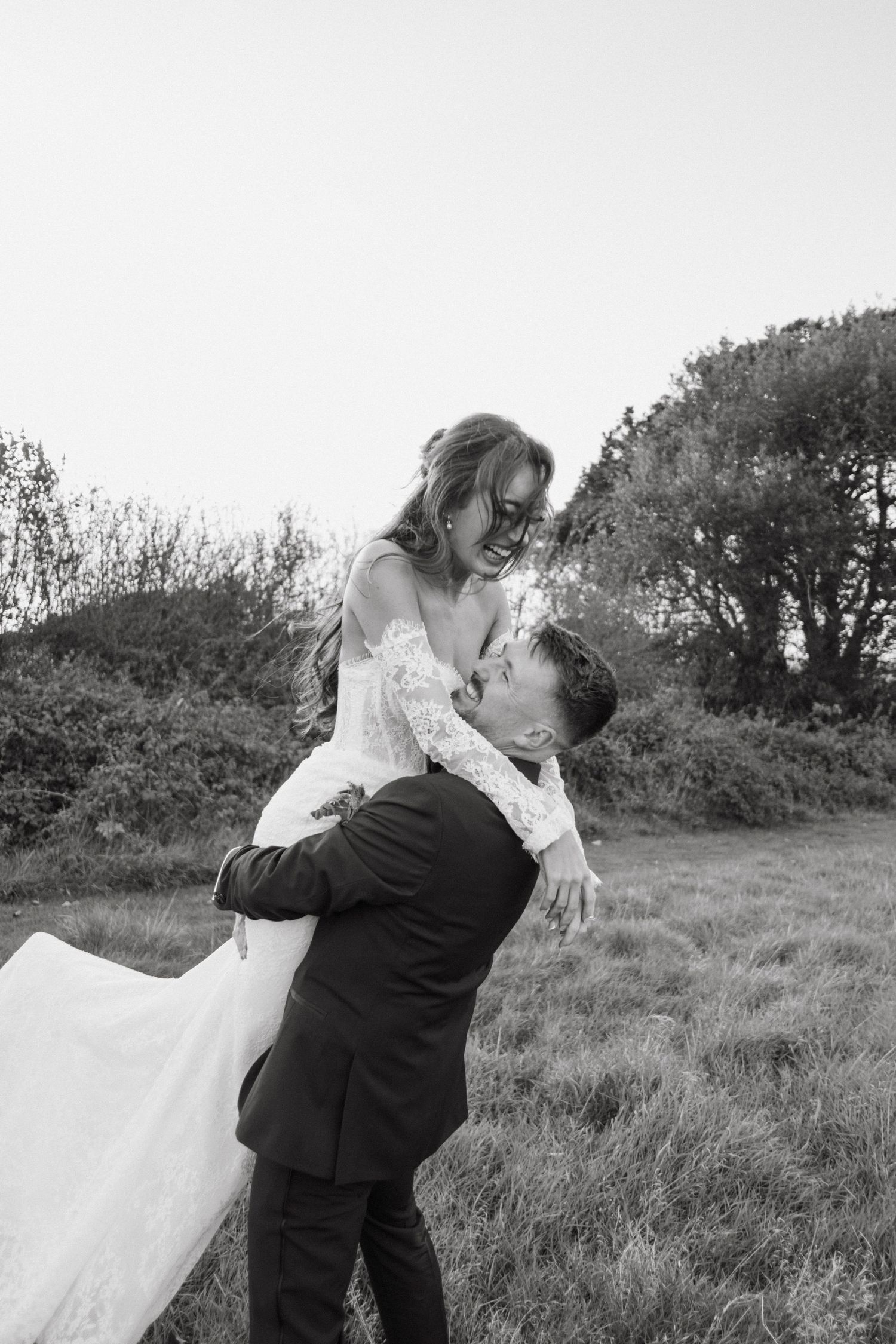 Groom lifts smiling bride in lace wedding dress outdoors in a black and white romantic wedding photo.
