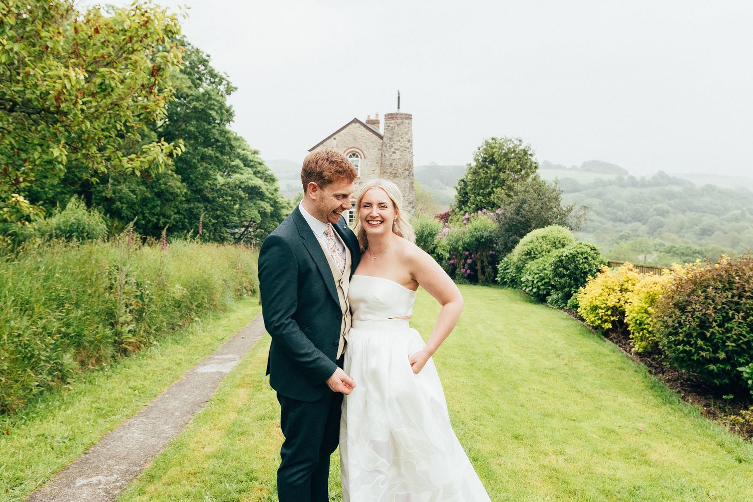 Smiling bride and groom pose together outside a stone church surrounded by lush green gardens.