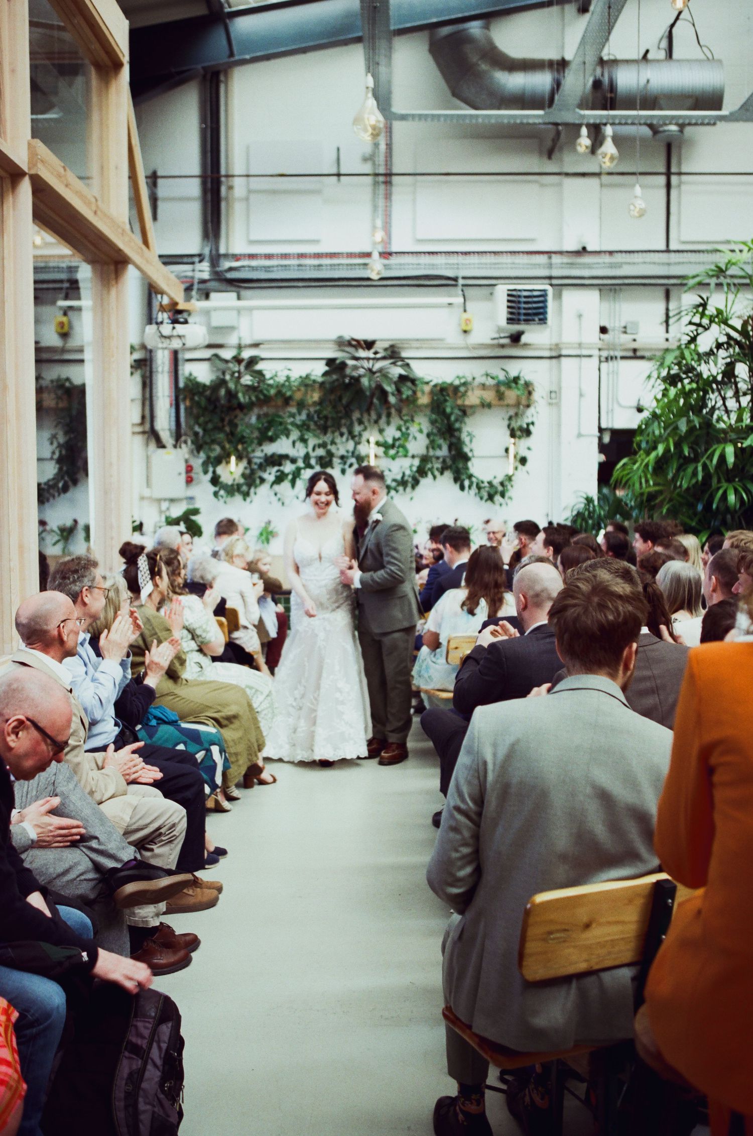 Wedding ceremony in industrial venue with couple at altar, seated guests, and lush green plant decorations.