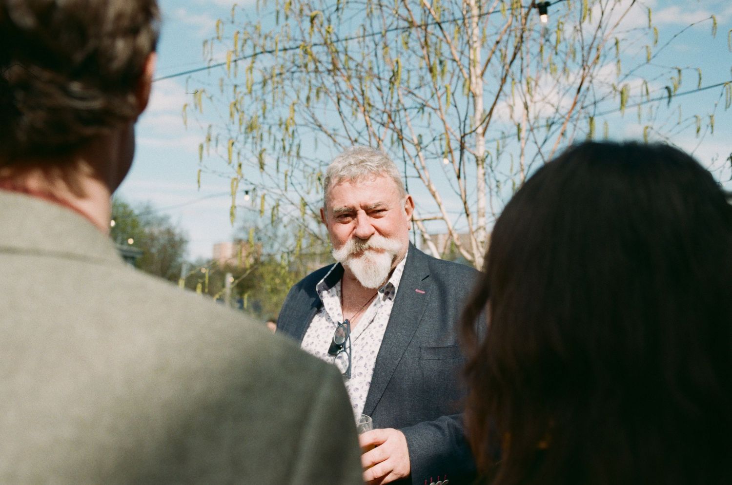 Older man with gray beard in blue blazer smiling outdoors, conversing with people near trees on a sunny day.
