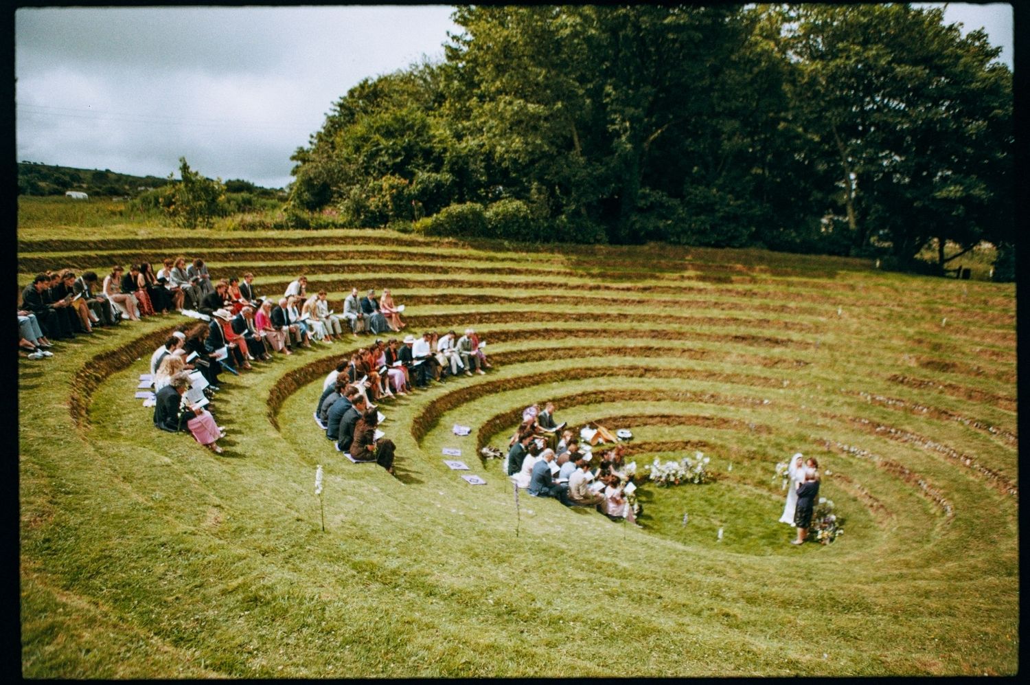 People gathered in an outdoor grass amphitheater with tiered seating, surrounded by trees on a cloudy day.