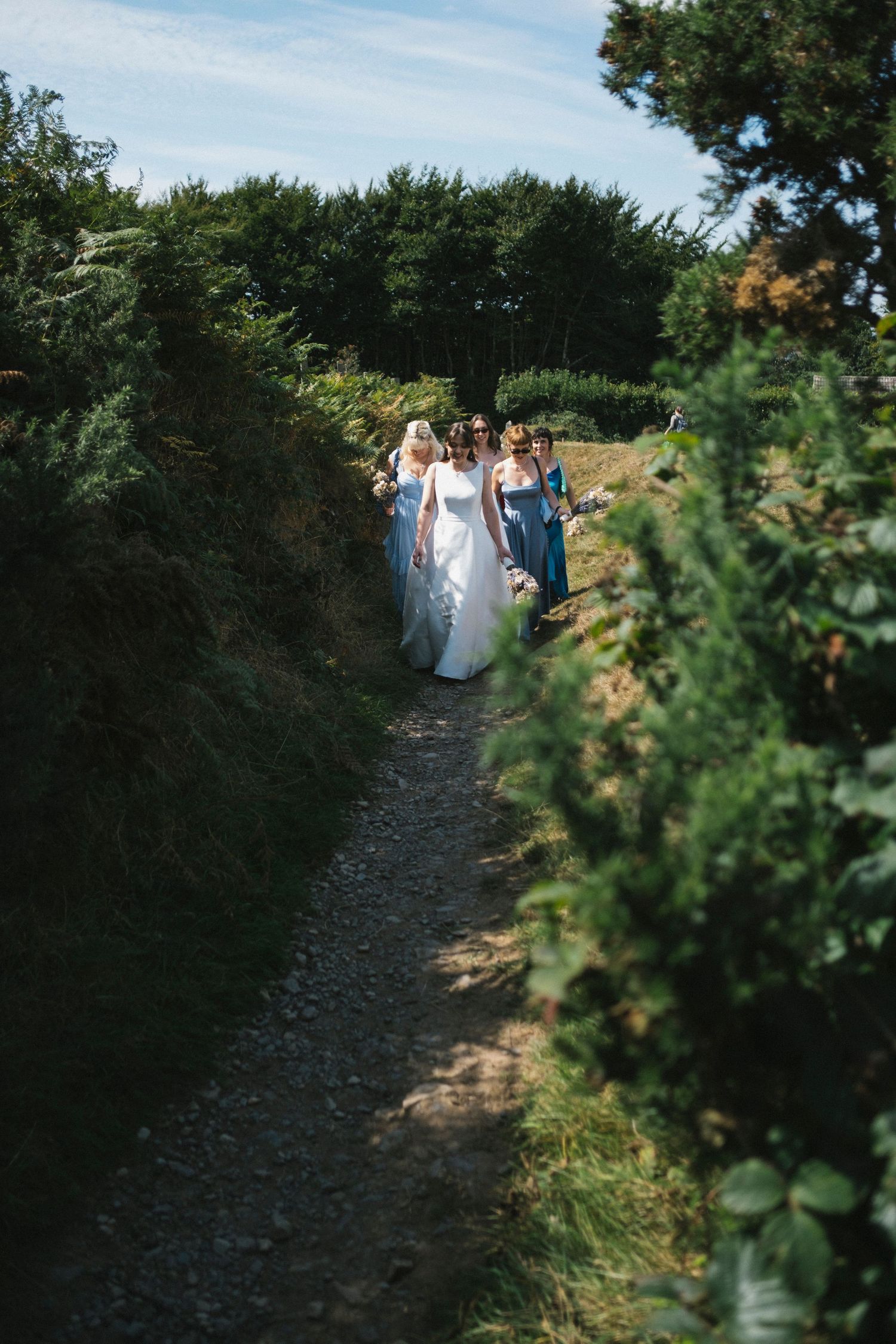 Bride and bridesmaids in blue gowns walk along a narrow sunken country path lined with greenery.