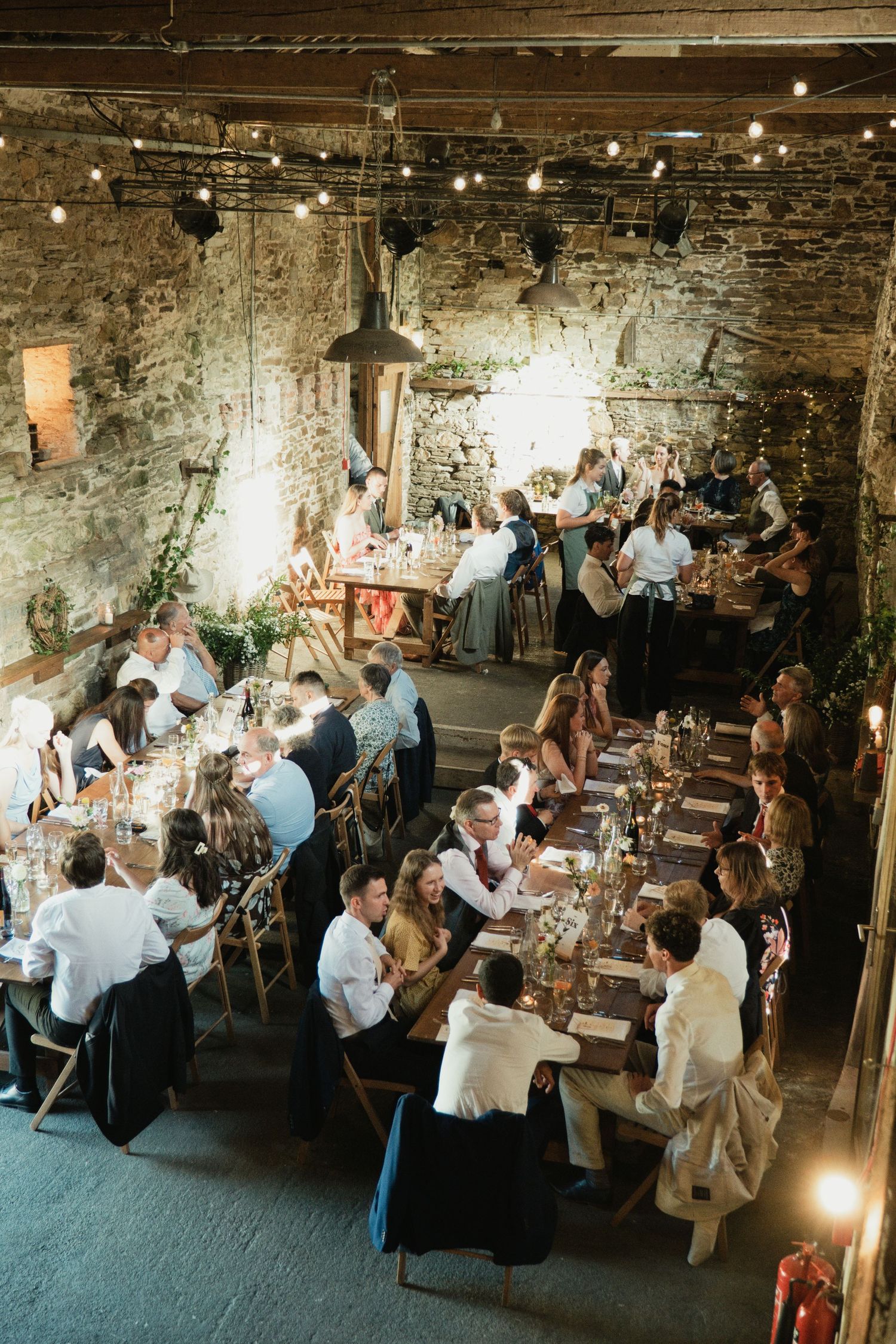 Guests dining at long wooden tables in a rustic stone barn wedding reception with string lights overhead.