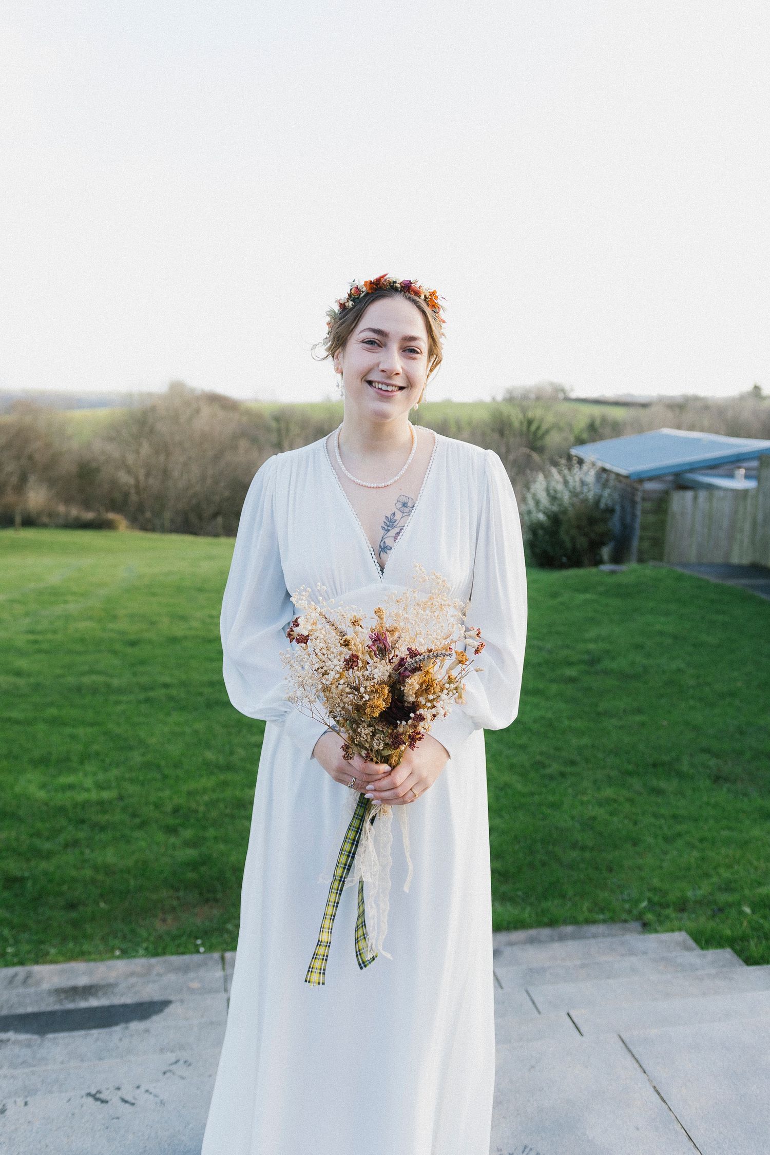 Smiling bride in a white dress holds a dried flower bouquet wearing a colorful floral crown.