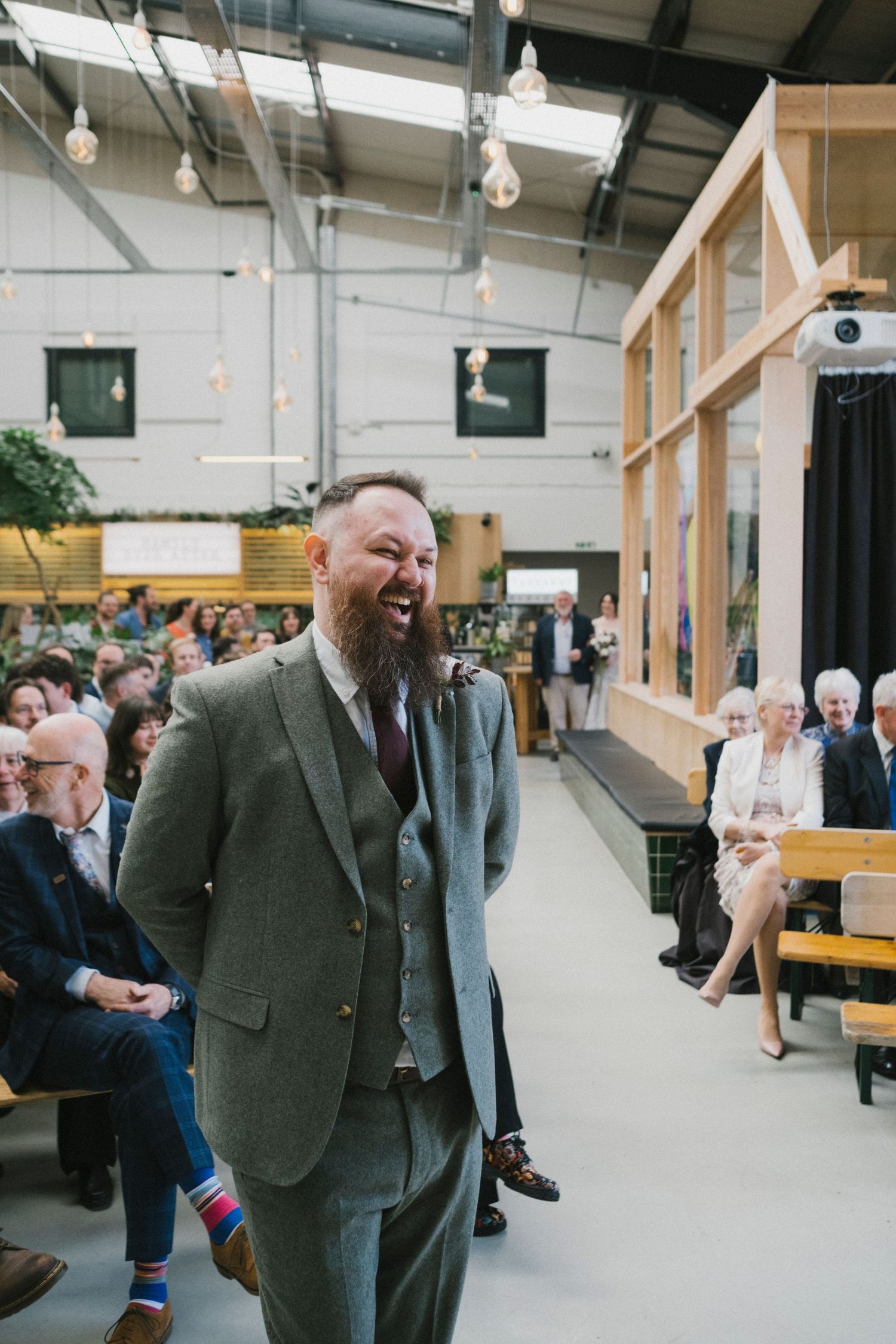 A laughing groom in a grey tweed suit stands at the altar during his wedding ceremony in an industrial venue.