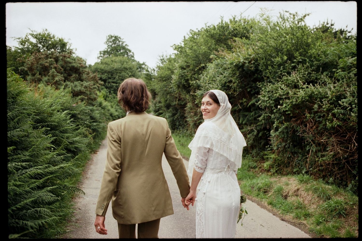 A bride in white dress and veil holds hands with groom in olive suit walking a tree-lined path.