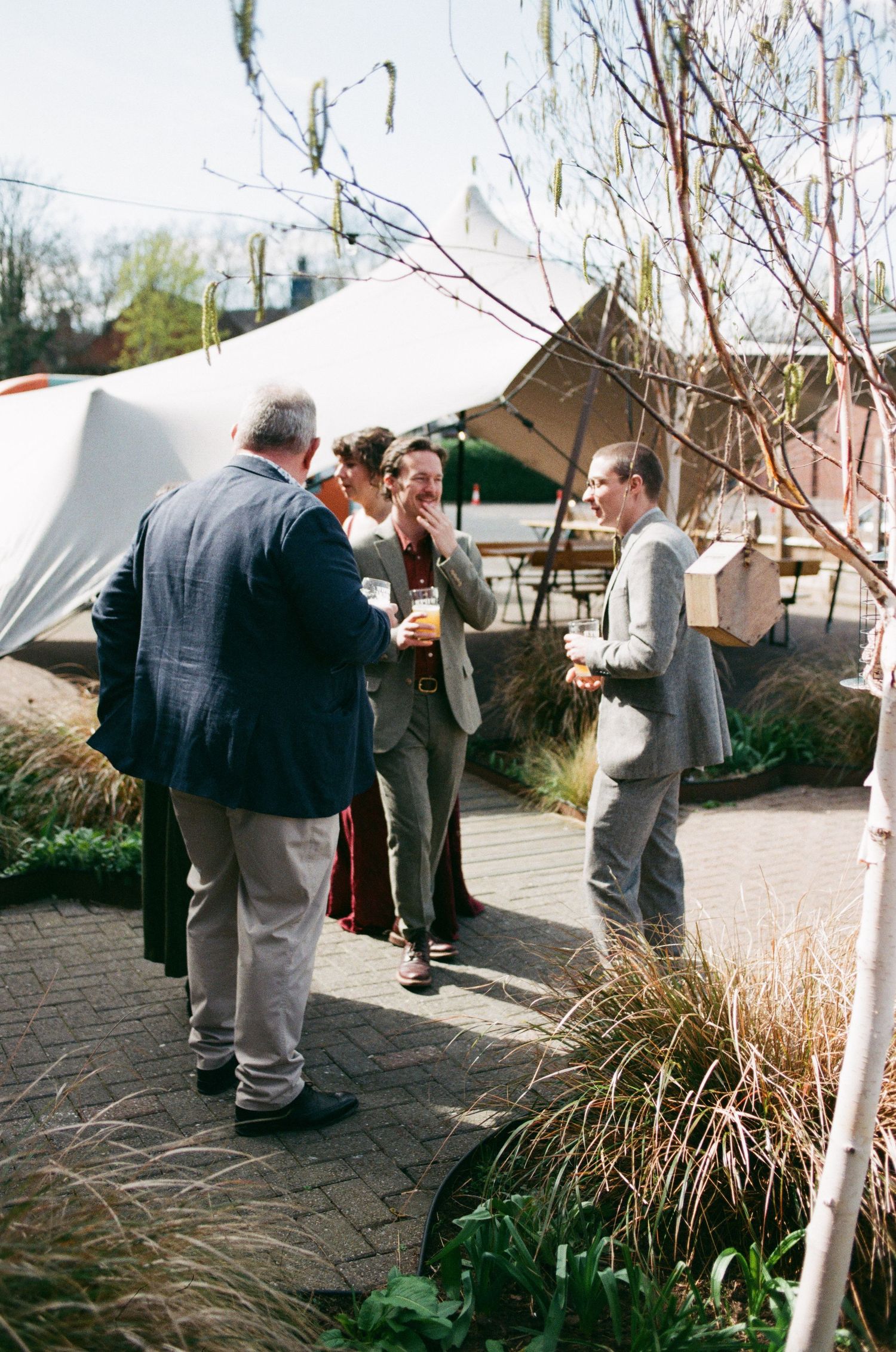 Men in suits socializing outdoors at a garden event with white tents and brick patio on a sunny day.