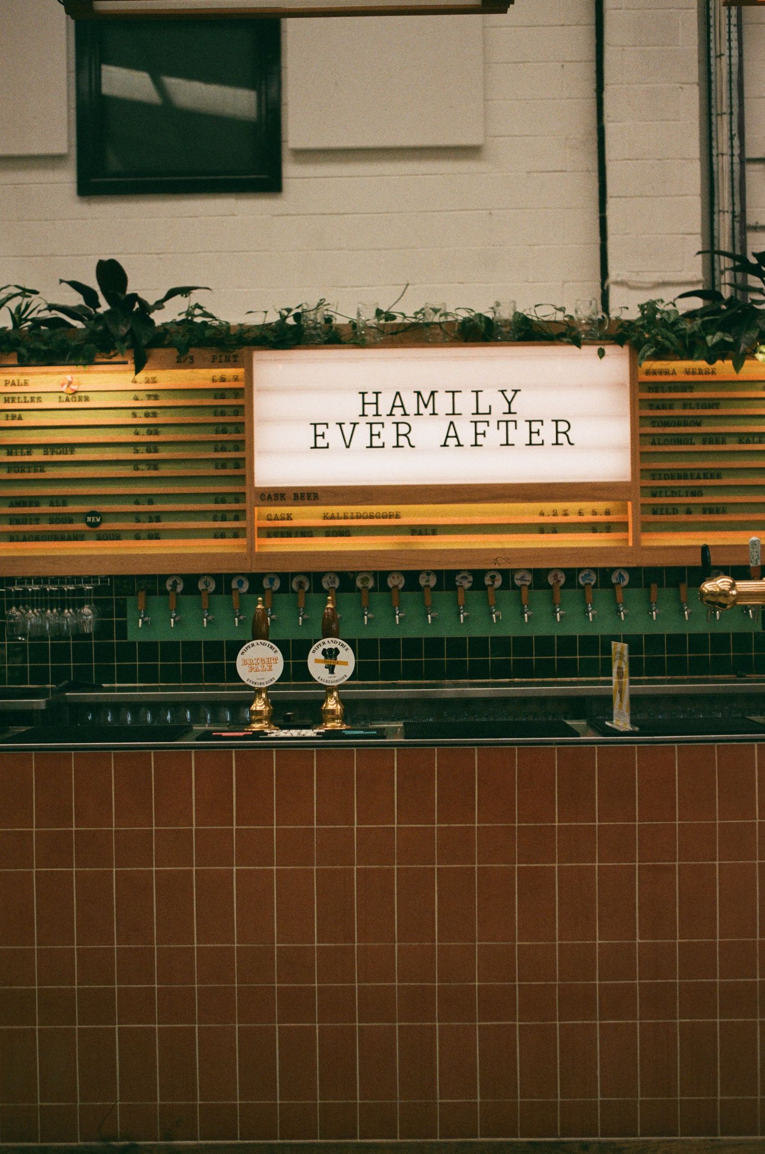Bar counter with illuminated sign reading Hamily Ever After, beer taps, green tiles, and menu boards with plants above.
