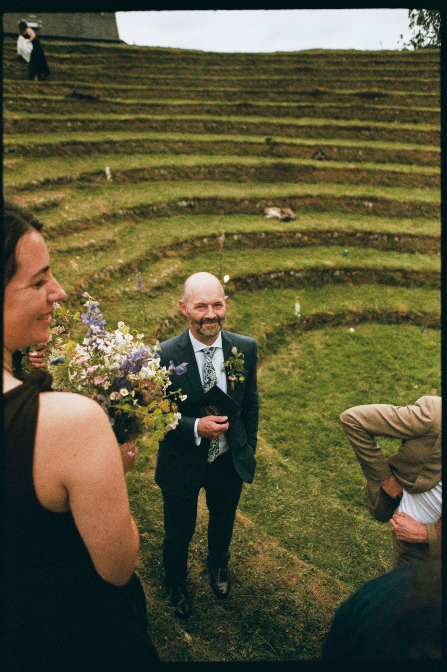 Smiling groom in a dark suit with floral boutonniere standing in a green terraced amphitheater at a wedding.