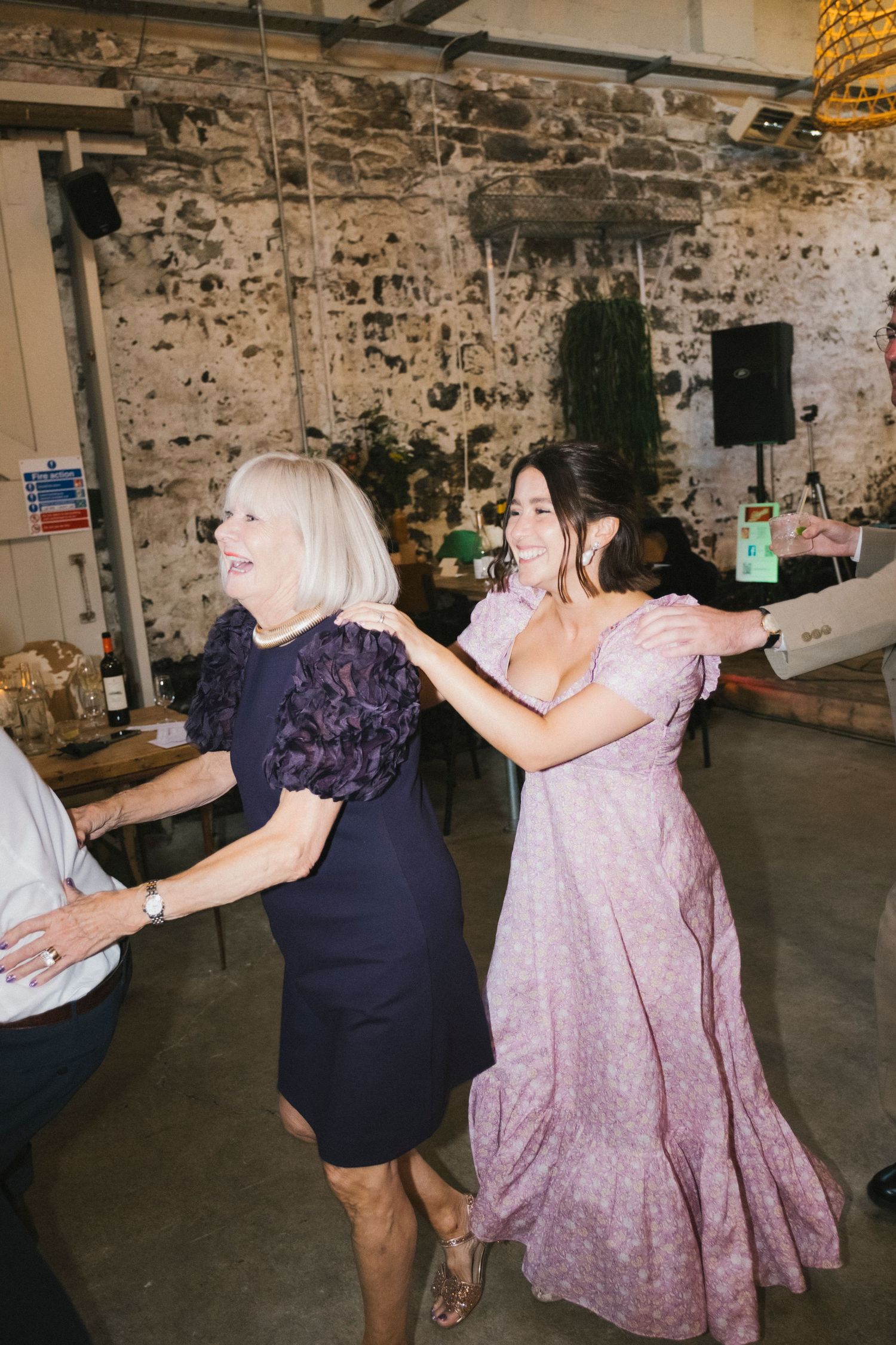 Two women dancing joyfully at a wedding reception in a rustic stone venue, wearing purple and pink dresses.