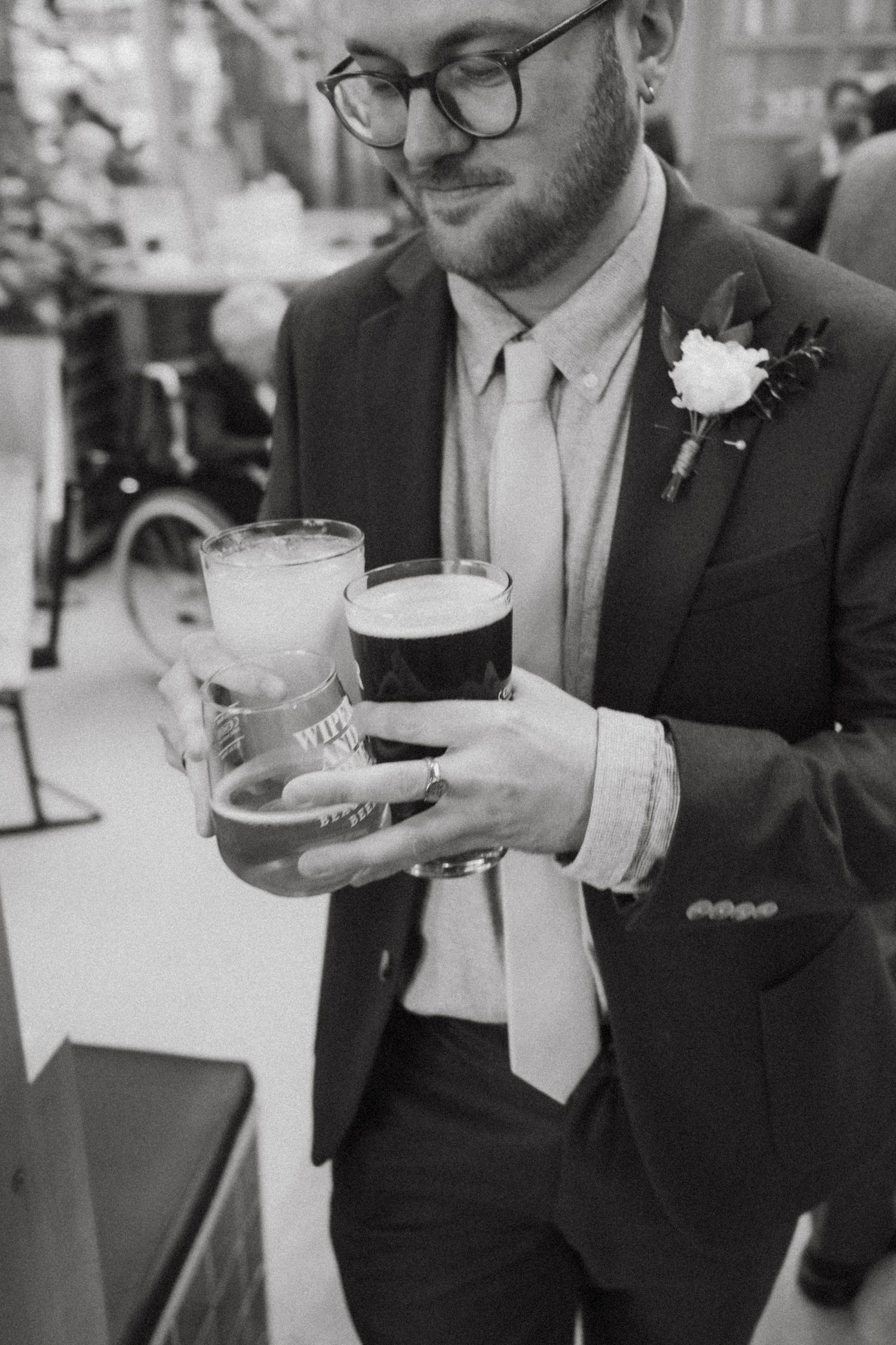 A groom in a suit with boutonniere holds multiple beer glasses at a wedding reception.