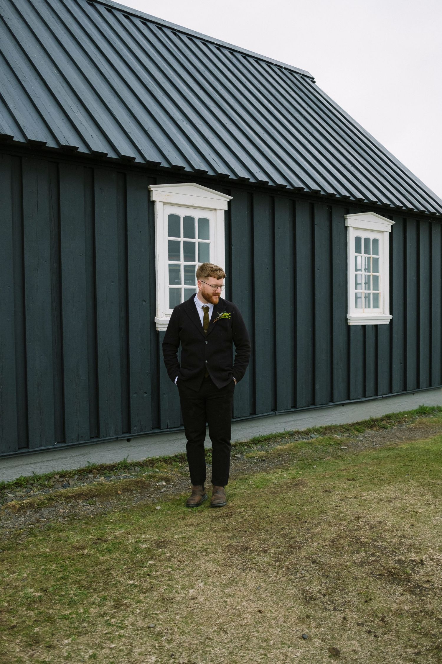 Groom in black suit standing outside a dark green wooden building in Iceland.