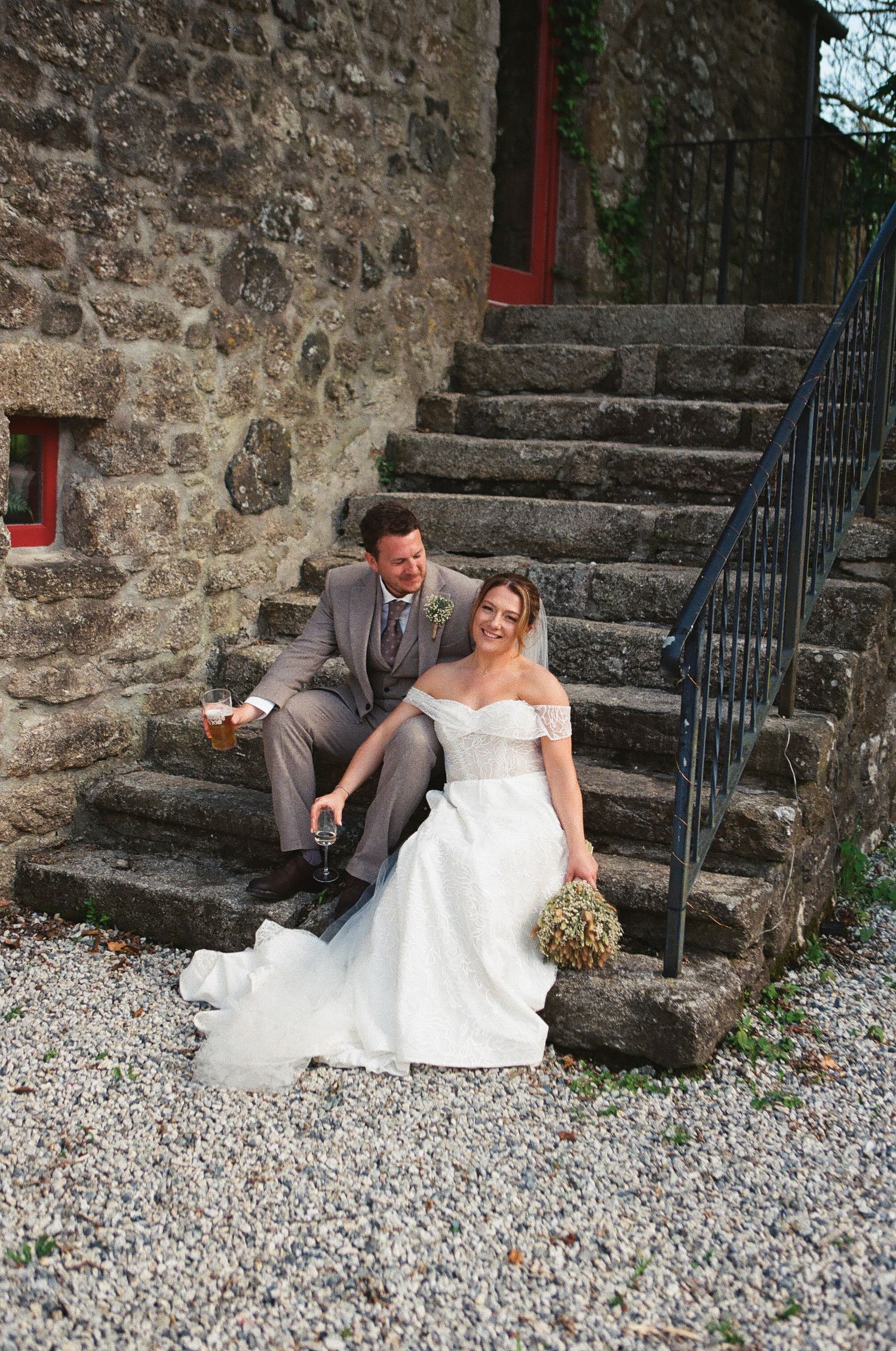 Bride and groom sit laughing together on ancient stone steps outside a rustic stone building on their wedding day.