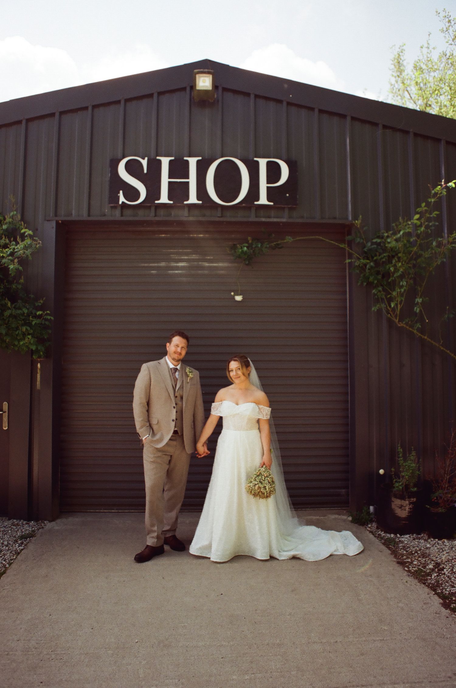 Bride and groom hold hands smiling in front of a dark metal SHOP building on their wedding day.