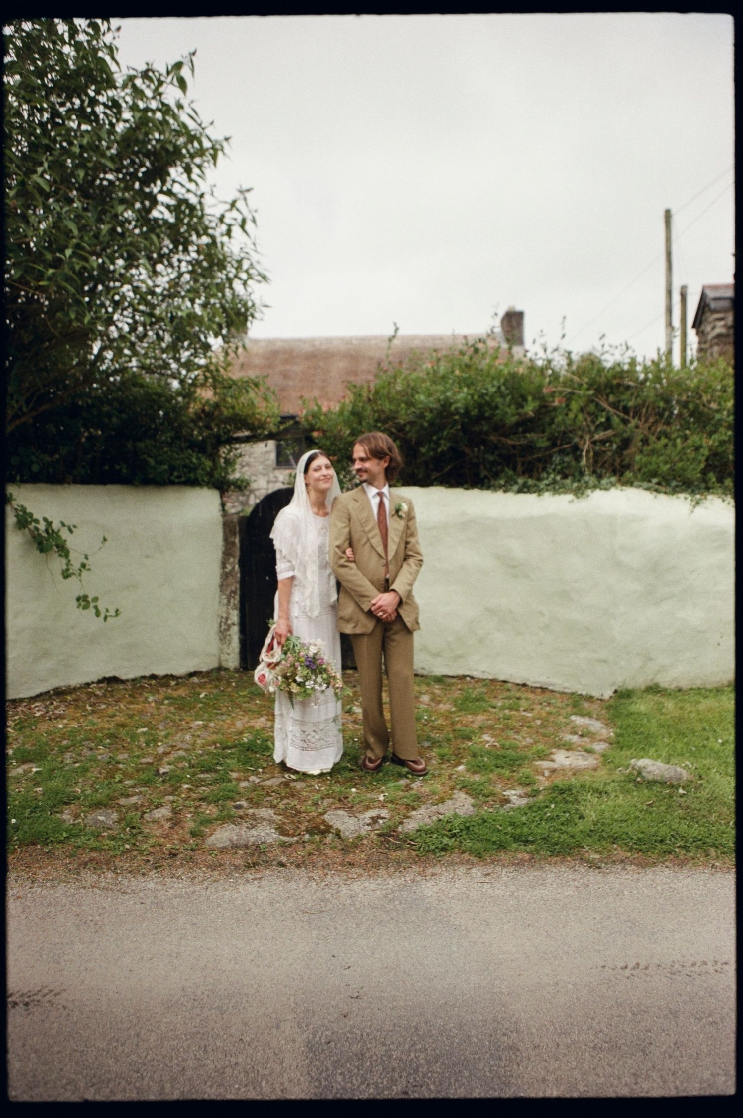A bride and groom pose together outdoors near a white stone wall, surrounded by greenery on their wedding day.