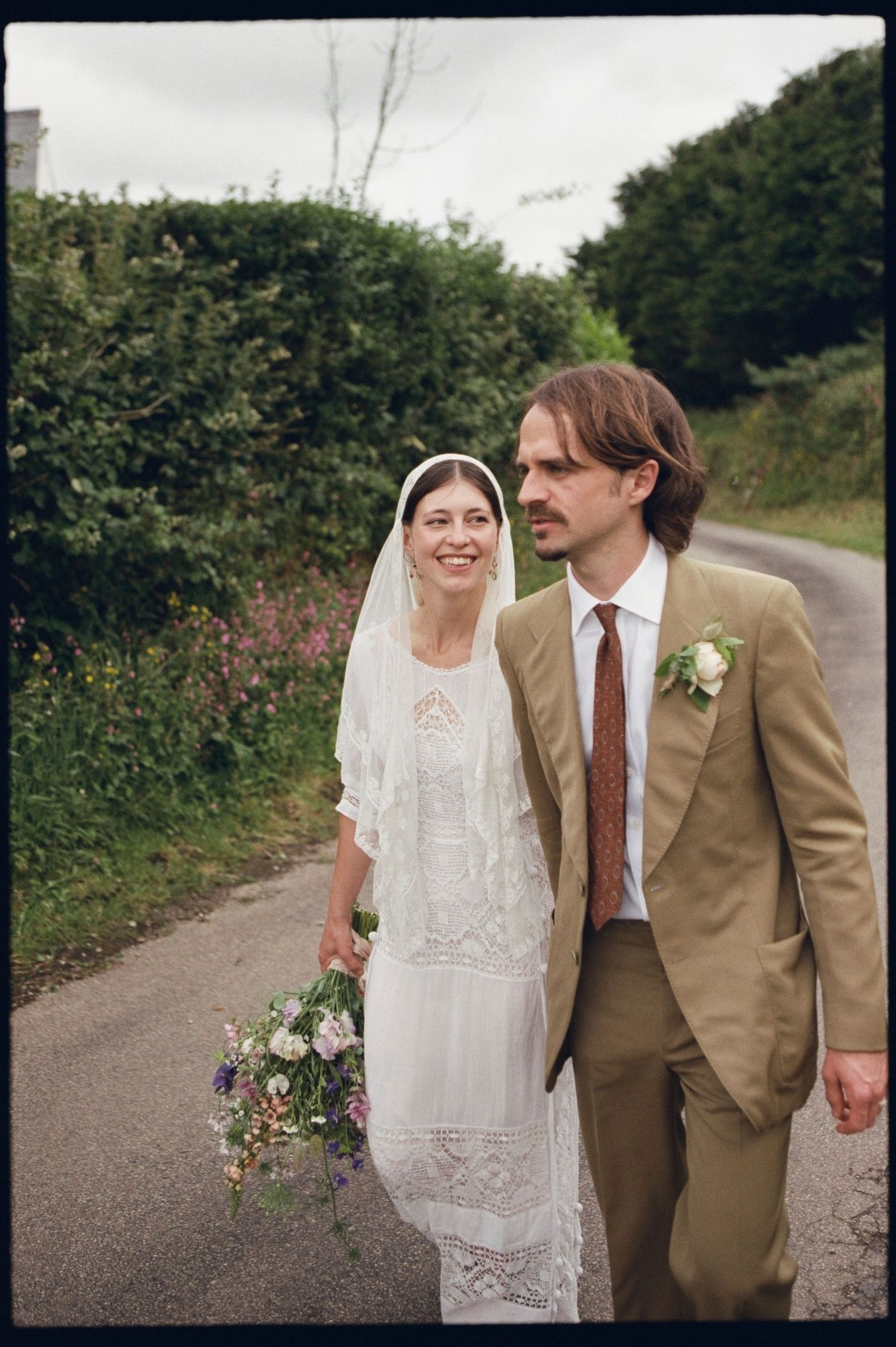 A bride in white lace dress and veil holds wildflowers beside a groom in tan suit on a country lane.