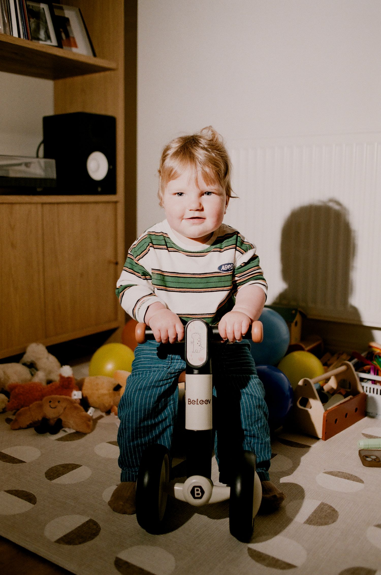 Adorable toddler riding a Balance bike toy in a sunny playroom full of toys.