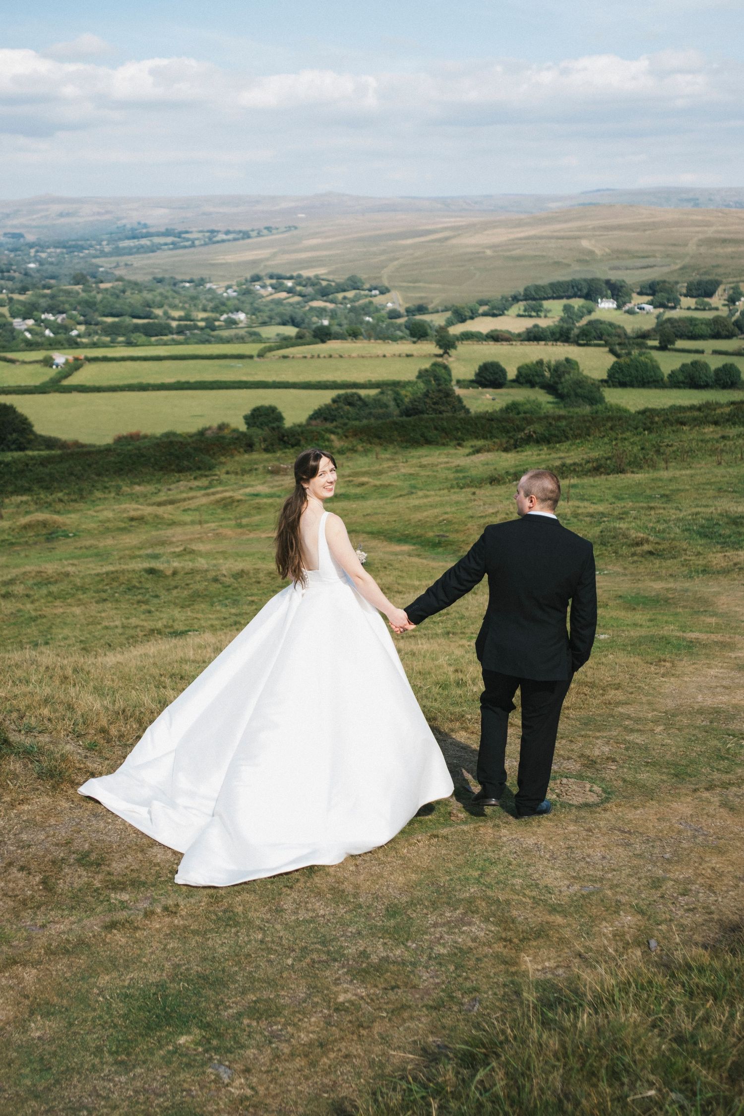 Bride in white gown and groom in black suit hold hands overlooking lush green countryside valley.