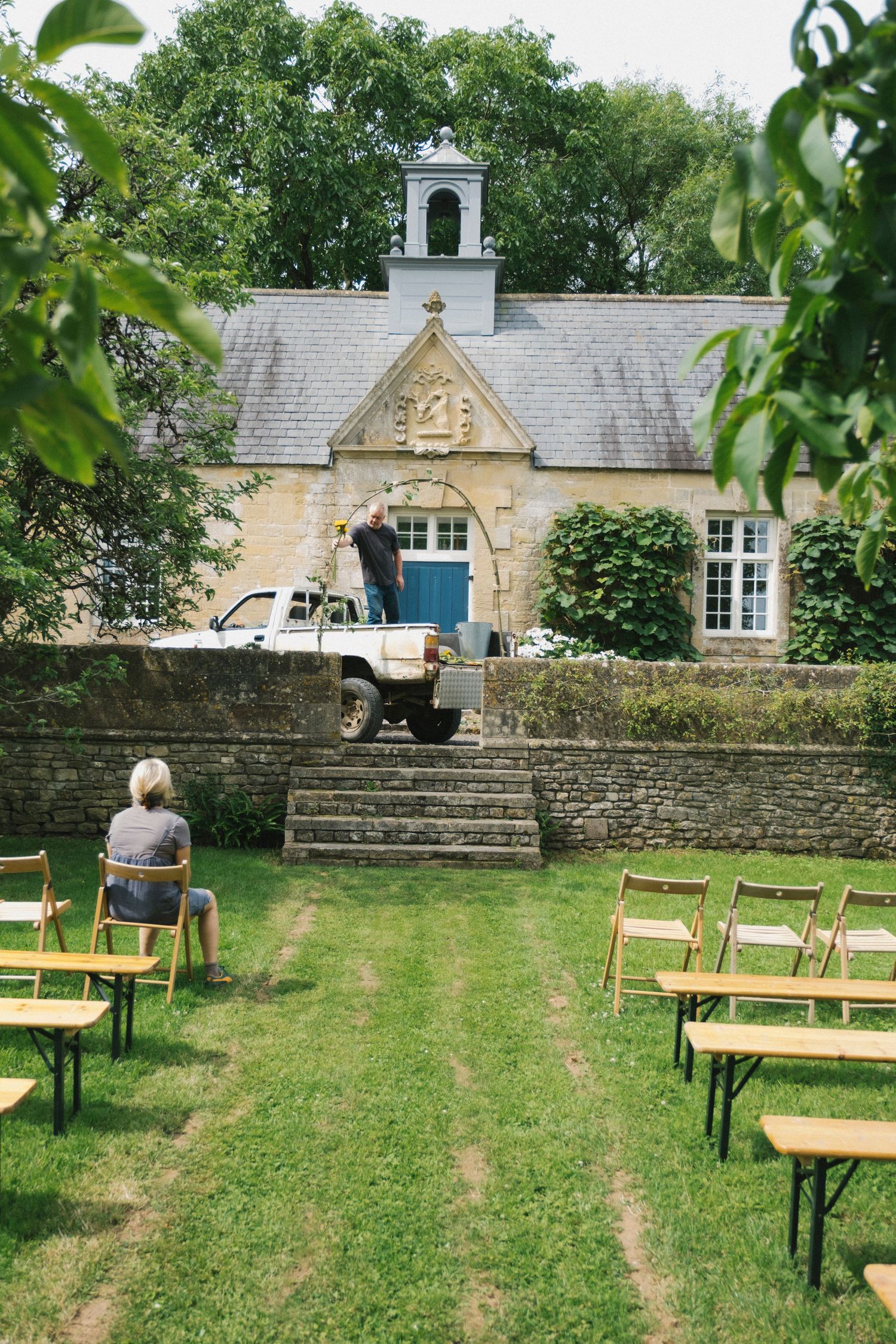 Outdoor garden ceremony setup with wooden chairs on grass facing a stone building with a bell tower.